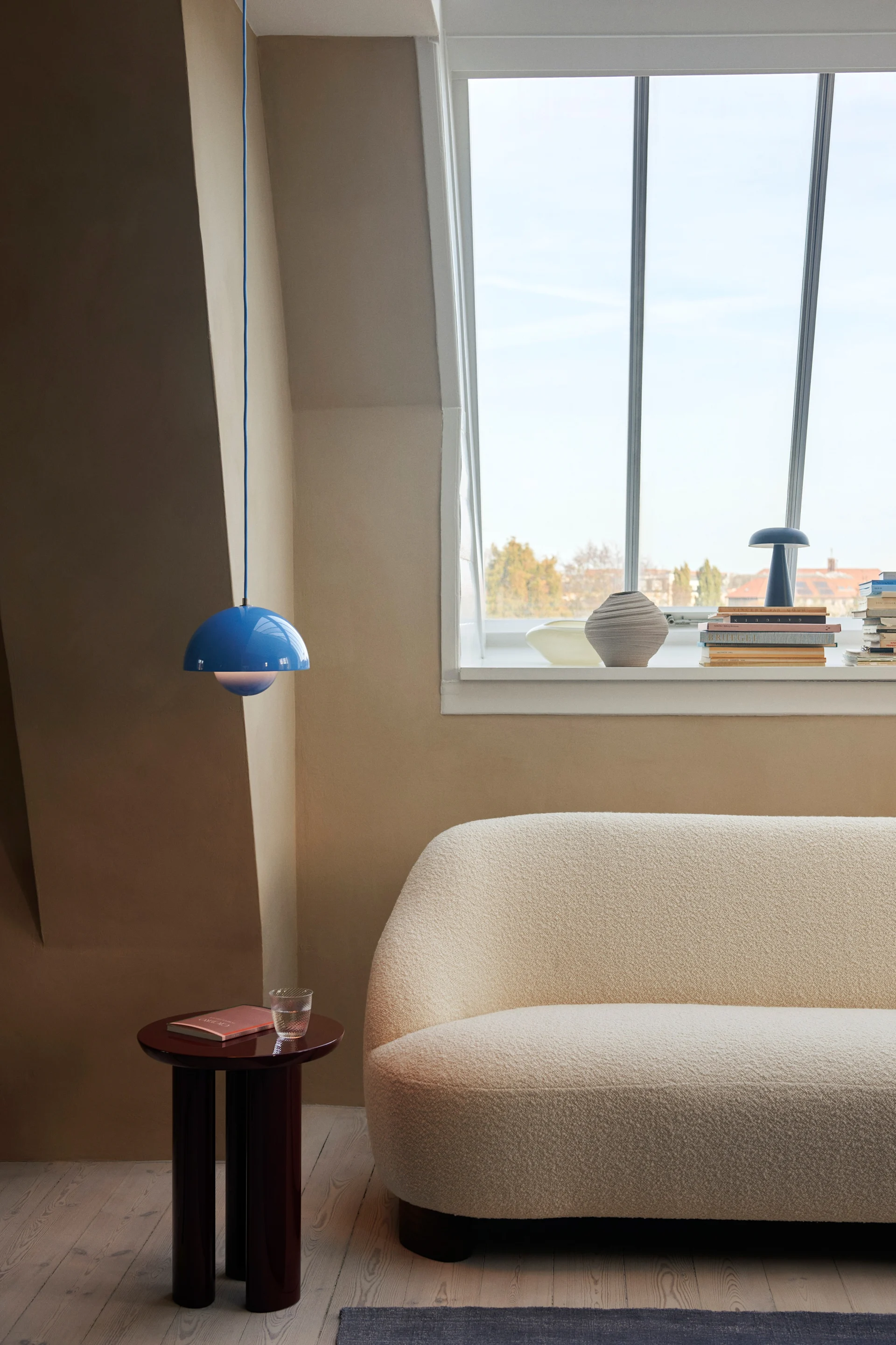 Flowerpot pendellampe VP1 i klar blå farge henger fra taket i et naturfarget rom, bildet viser også et sidebord i mørkt tre og en sofa i naturfarge.