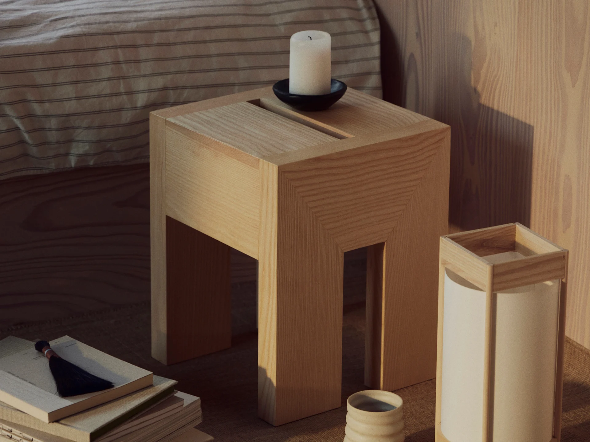 Et sidebord i tre med et hvitt lys i en svart holder, en stabel bøker med en dusk og en lampe med en keramisk kopp.