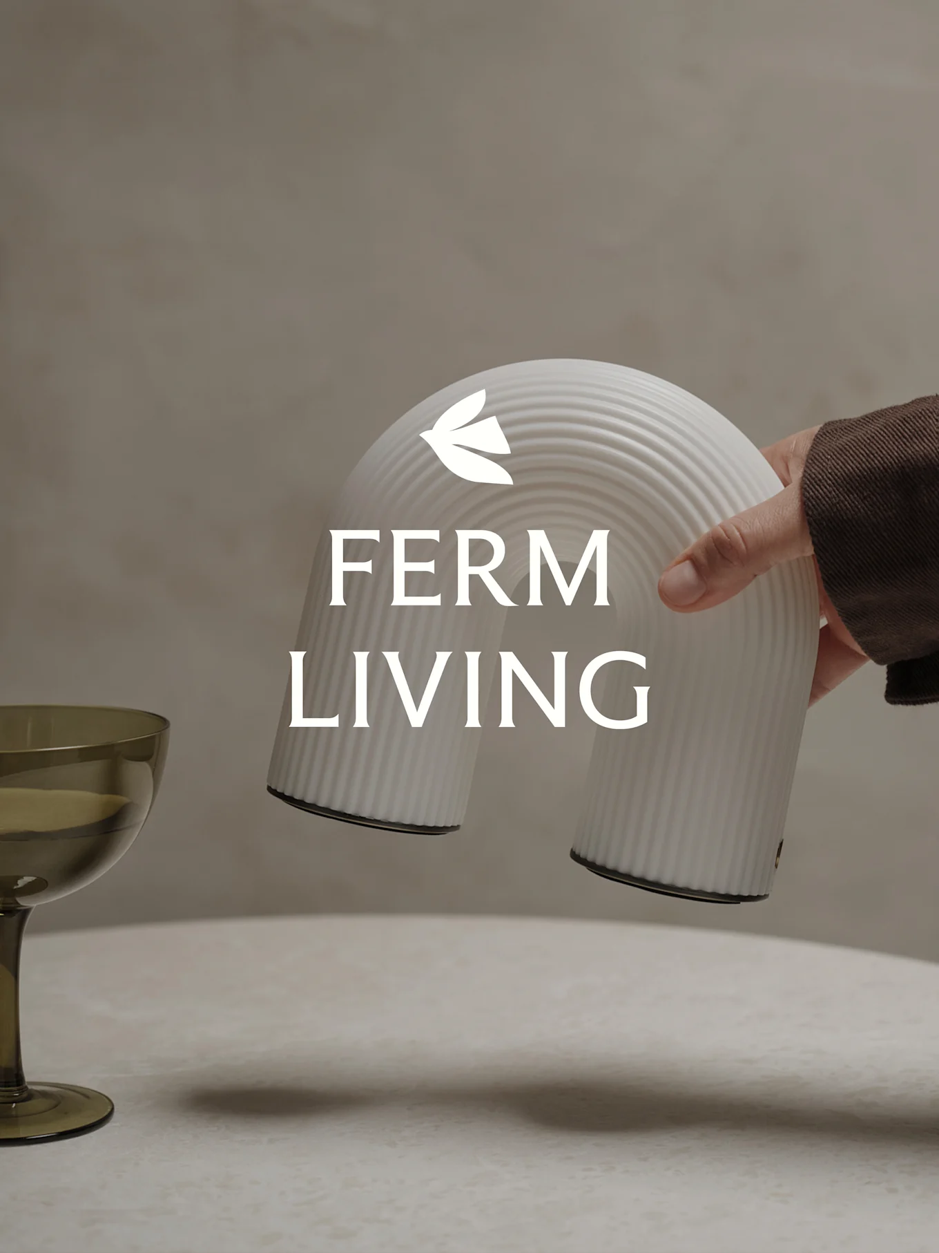 Hånd holder et hvitt ribbet bueformet dekorativt objekt, med et grønt glass i nærheten.