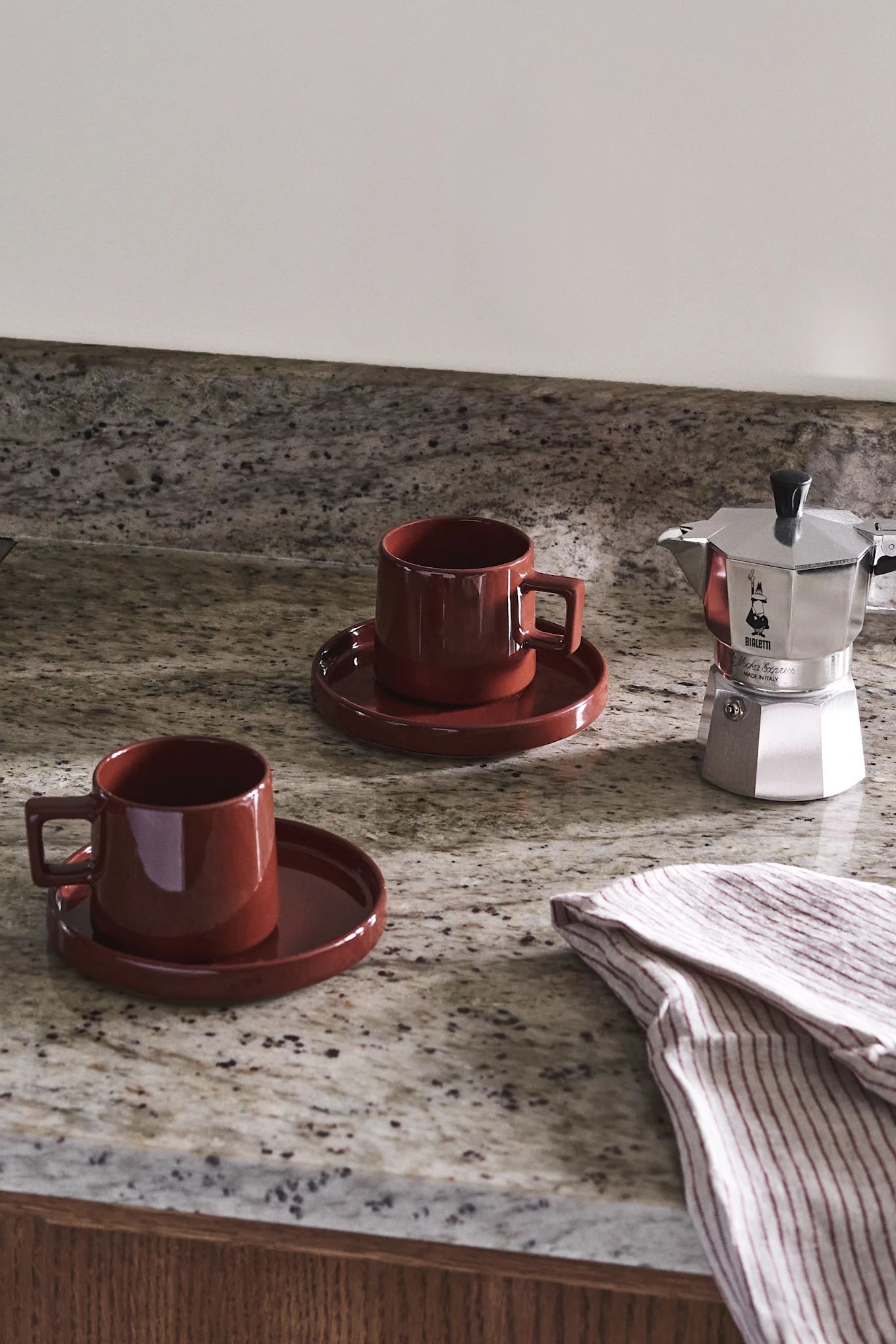 To røde keramiske krus og en sølvkaffemaskin på en flekkete granittbenkeplate med et stripet håndkle.