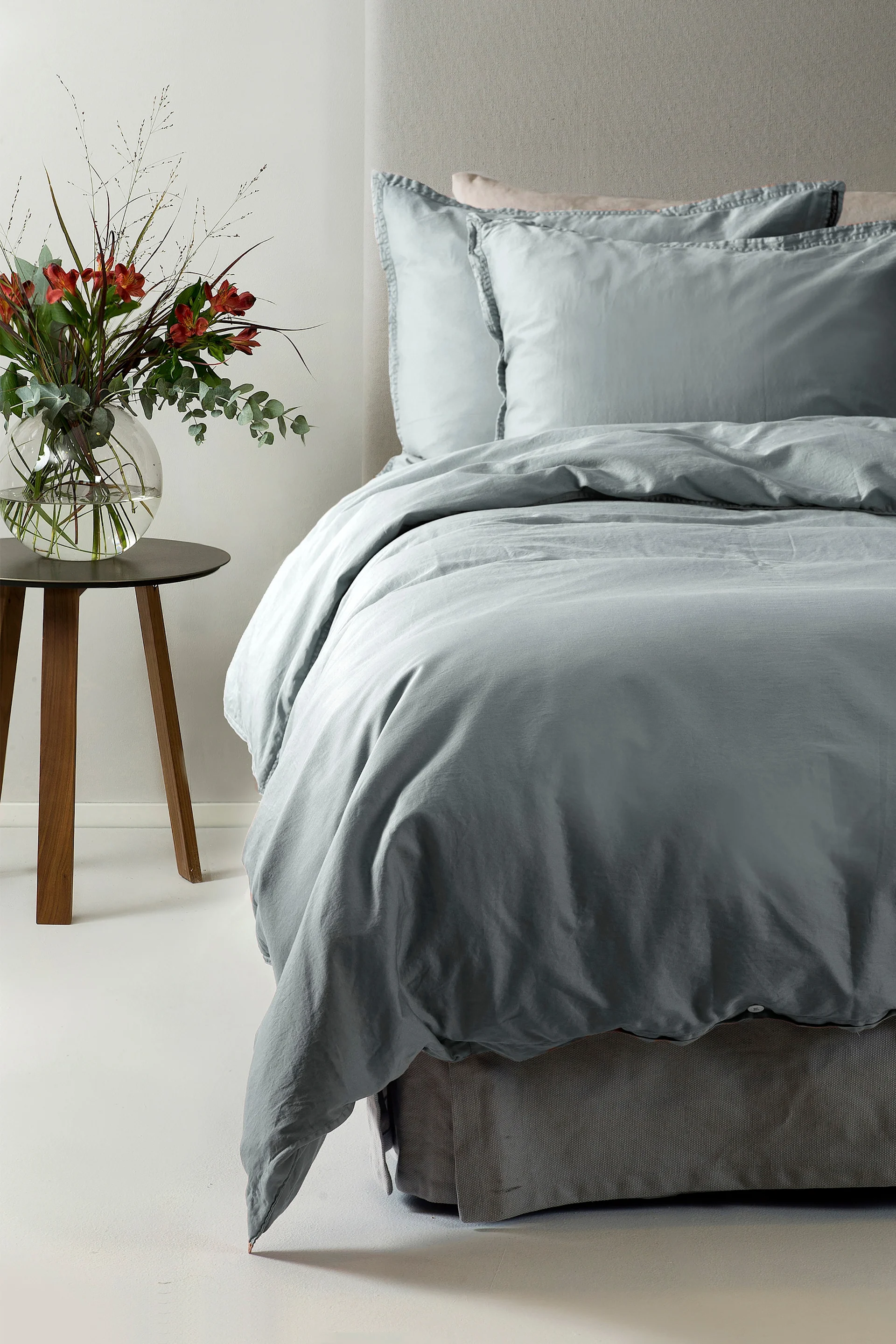 En seng med lyseblått sengetøy og to puter, ved siden av et nattbord i tre med en vase med røde blomster.