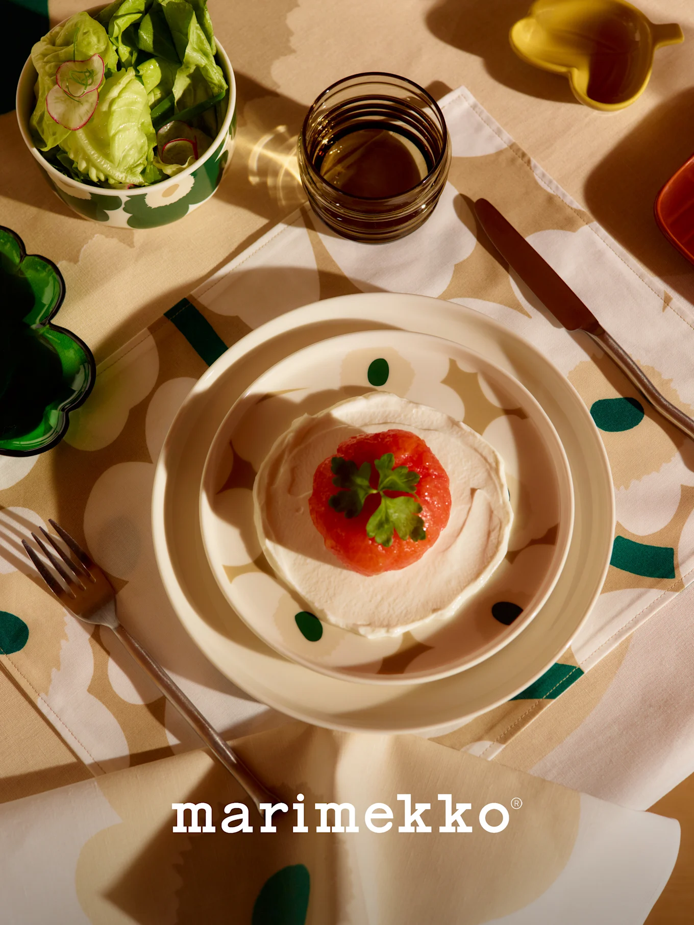 Marimekko borddekking med mønstret salatbolle, rett med rød frukt og krem, glass og bestikk på en blomstrete duk.