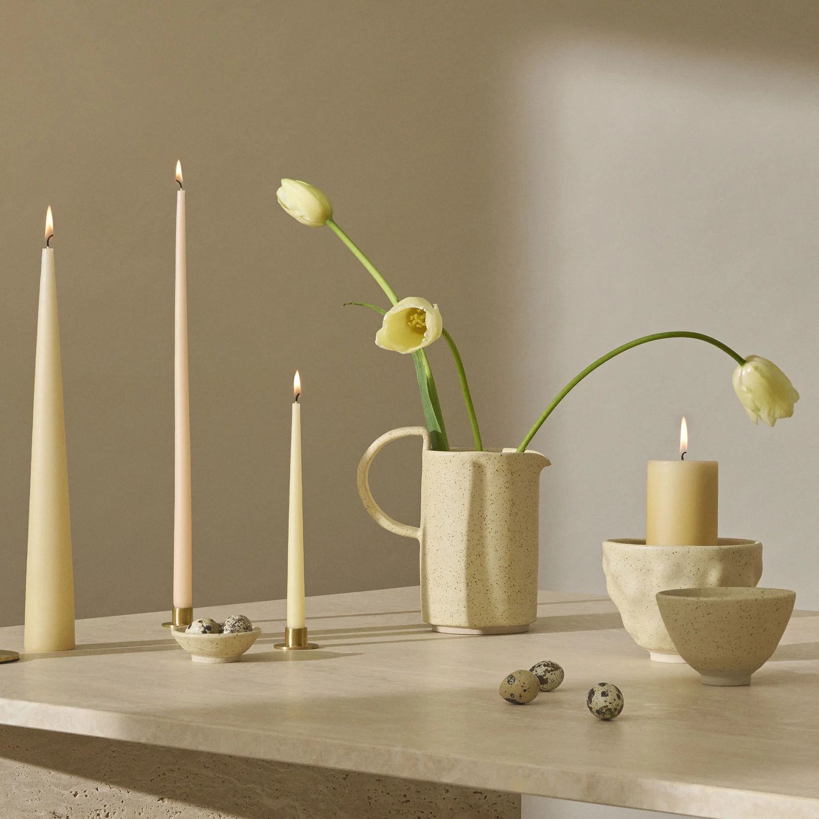 Et stilleben med tente stearinlys, blekgule tulipaner i en spraglete mugge og vaktel-egg på en teksturert overflate.