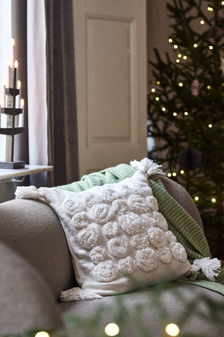 En hvit pute med dusker og et grønt pledd på sofaen for en moderne og minimalistisk julepynt i år.