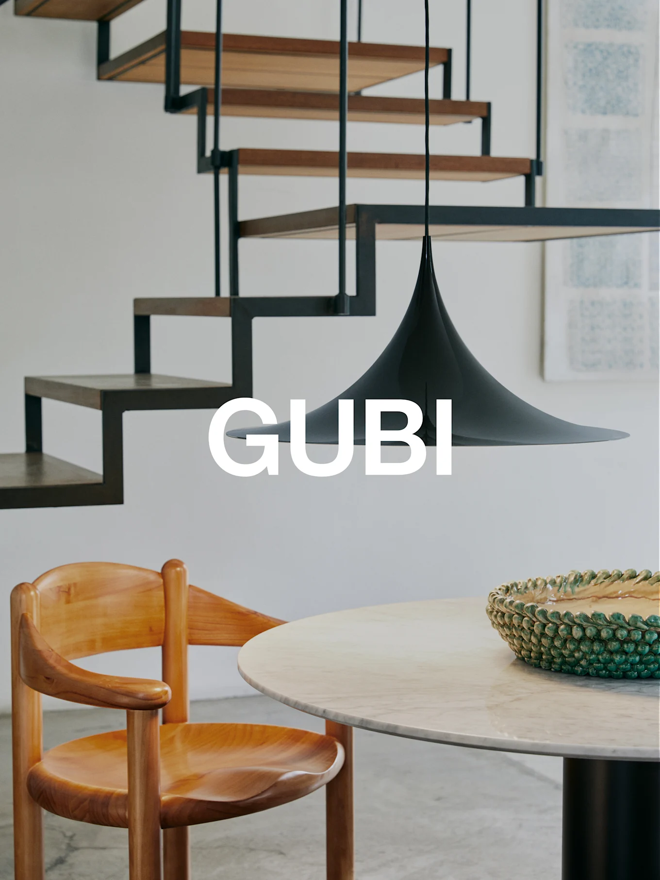 GUBI-logo i et moderne interiør med trestol, marmorbord, grønn bolle, svart pendellampe og en moderne trapp.