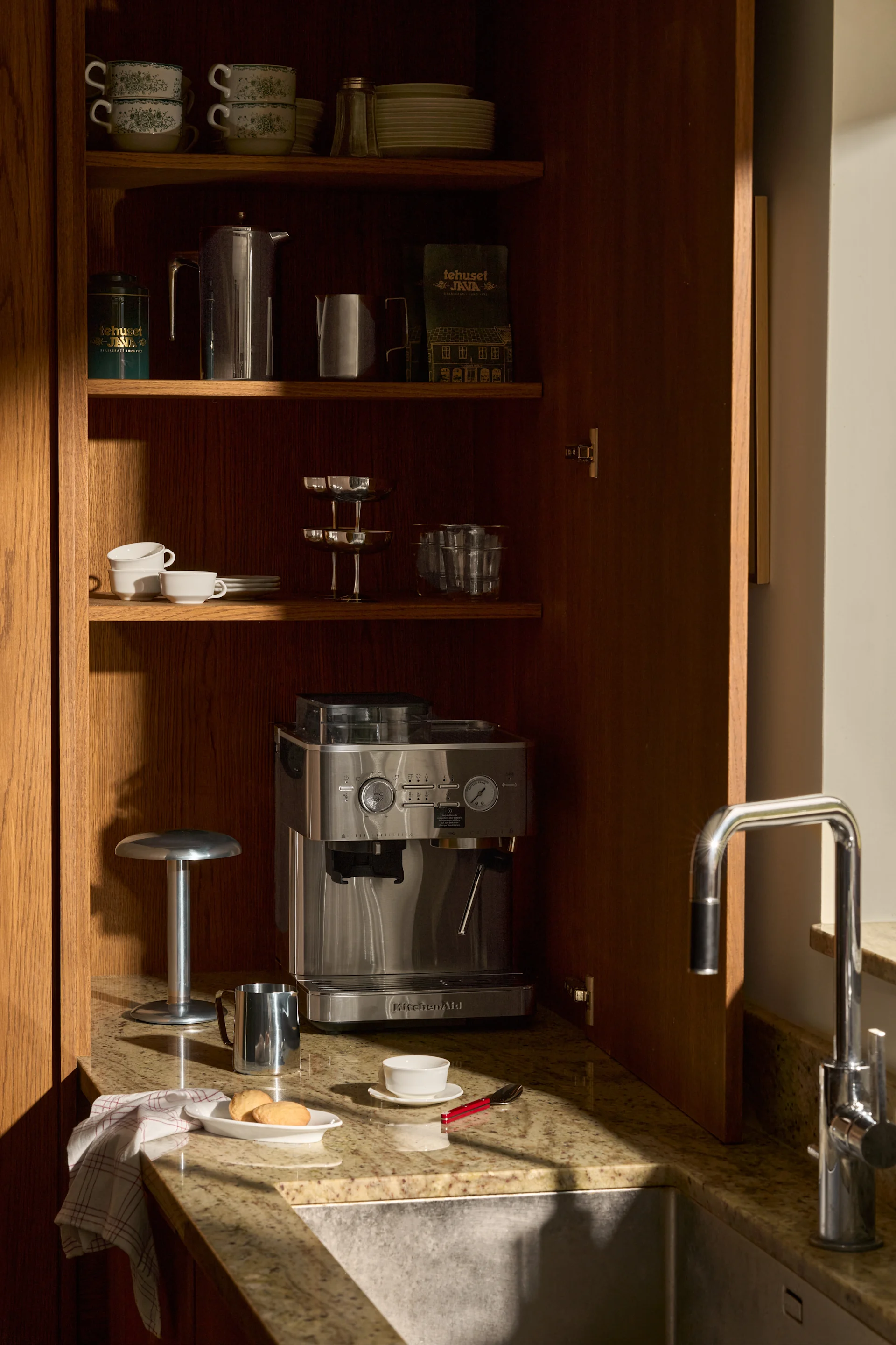 Åpent treskap med KitchenAid espressomaskin, stablede kopper, tallerkener, kaffeting og vask på en granittbenkeplate.