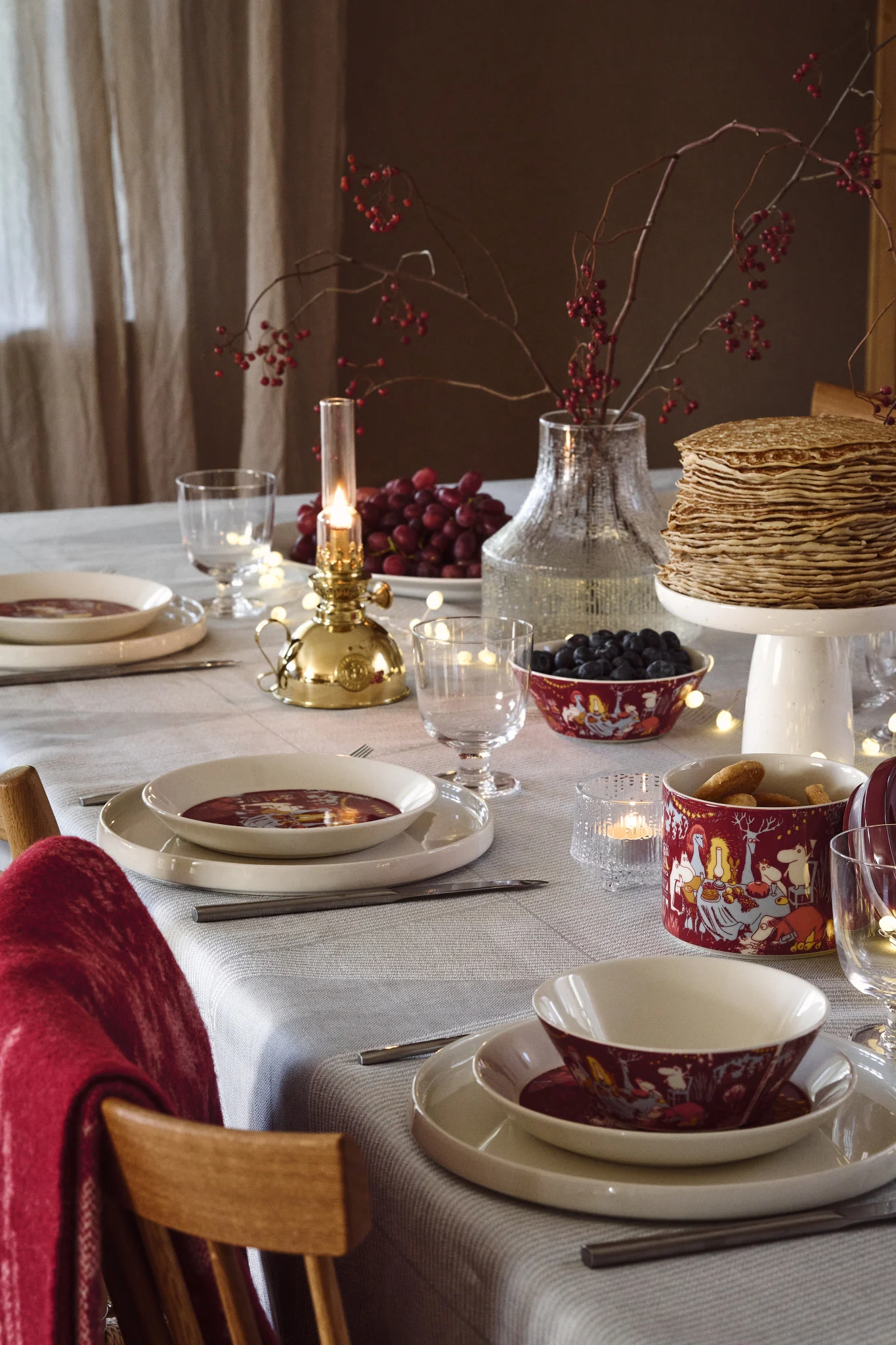 En leken borddekking jul 2025 skapes med rødt servise pyntet med Mummi-motiv, kombinert med fargerike bær som gir en varm og innbydende stemning på bordet.
