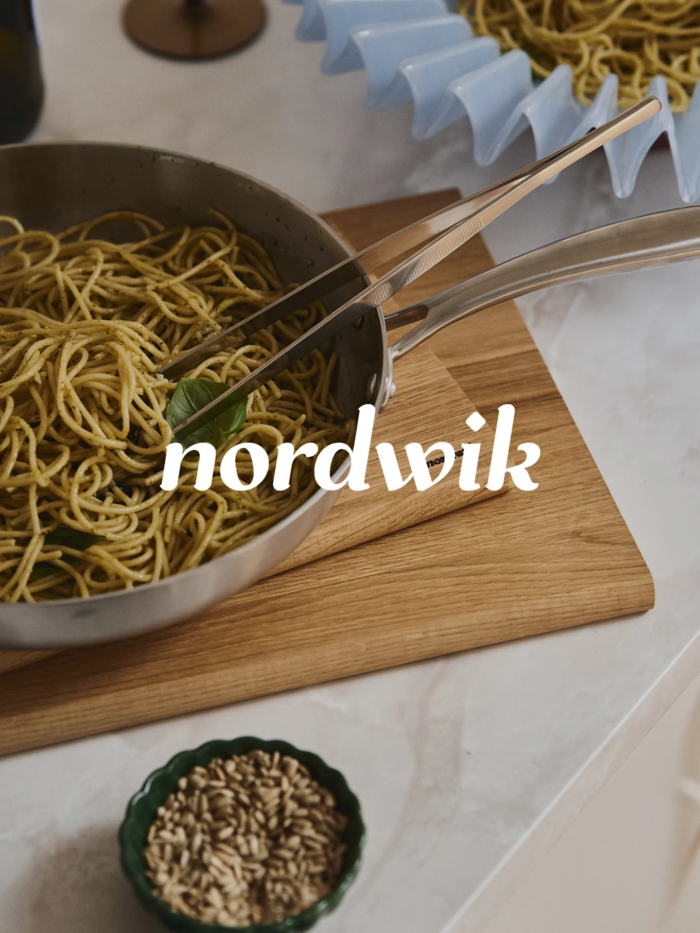 Spaghetti med pesto i en stålkasserolle med tang på et skjærebrett i tre. En skål med frø under, og et pastastativ i bakgrunnen. 'nordwik' tekstoverlegg.