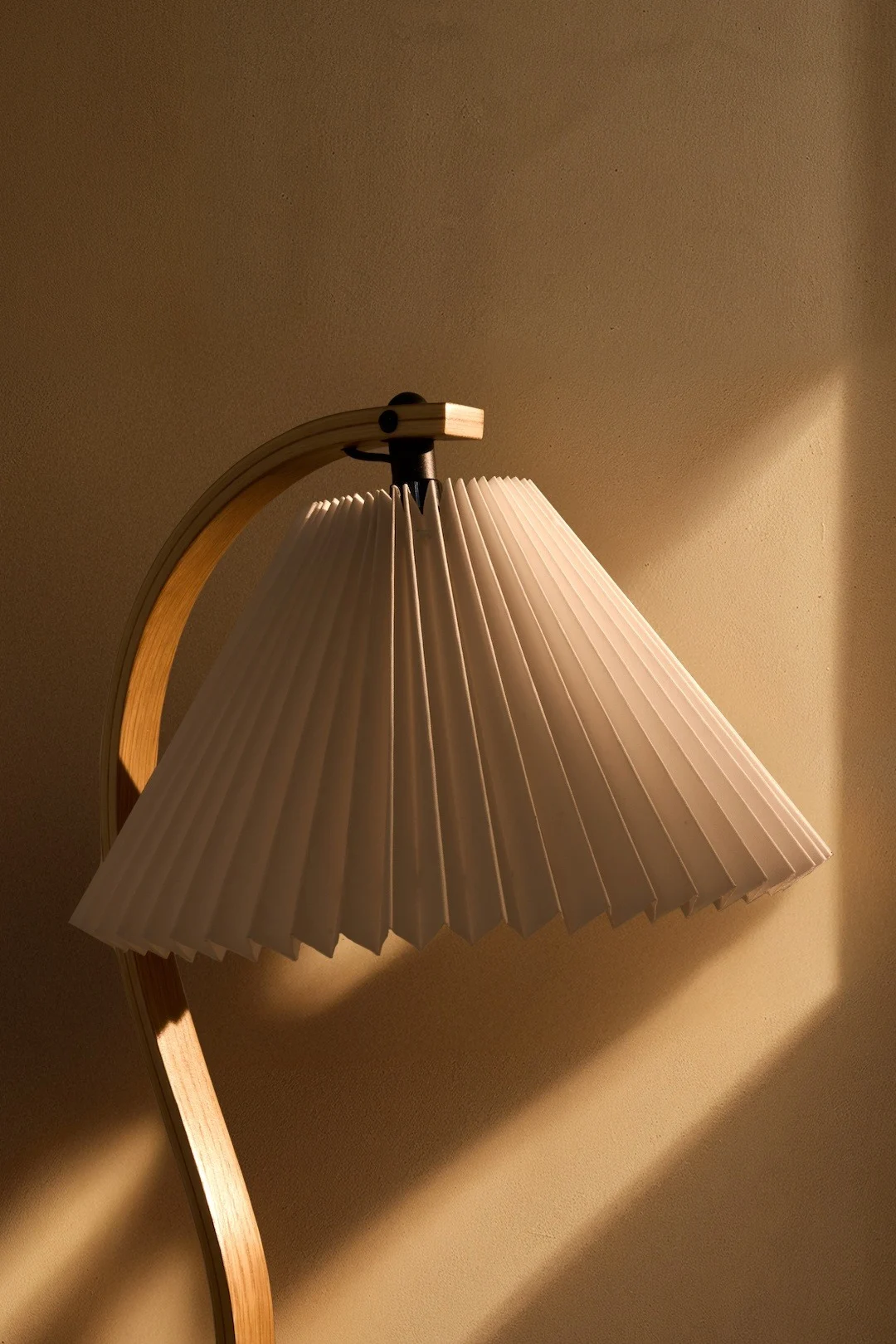 En lampe med en hvit plissert skjerm og en buet trebunn, som kaster varmt lys på en teksturert vegg.