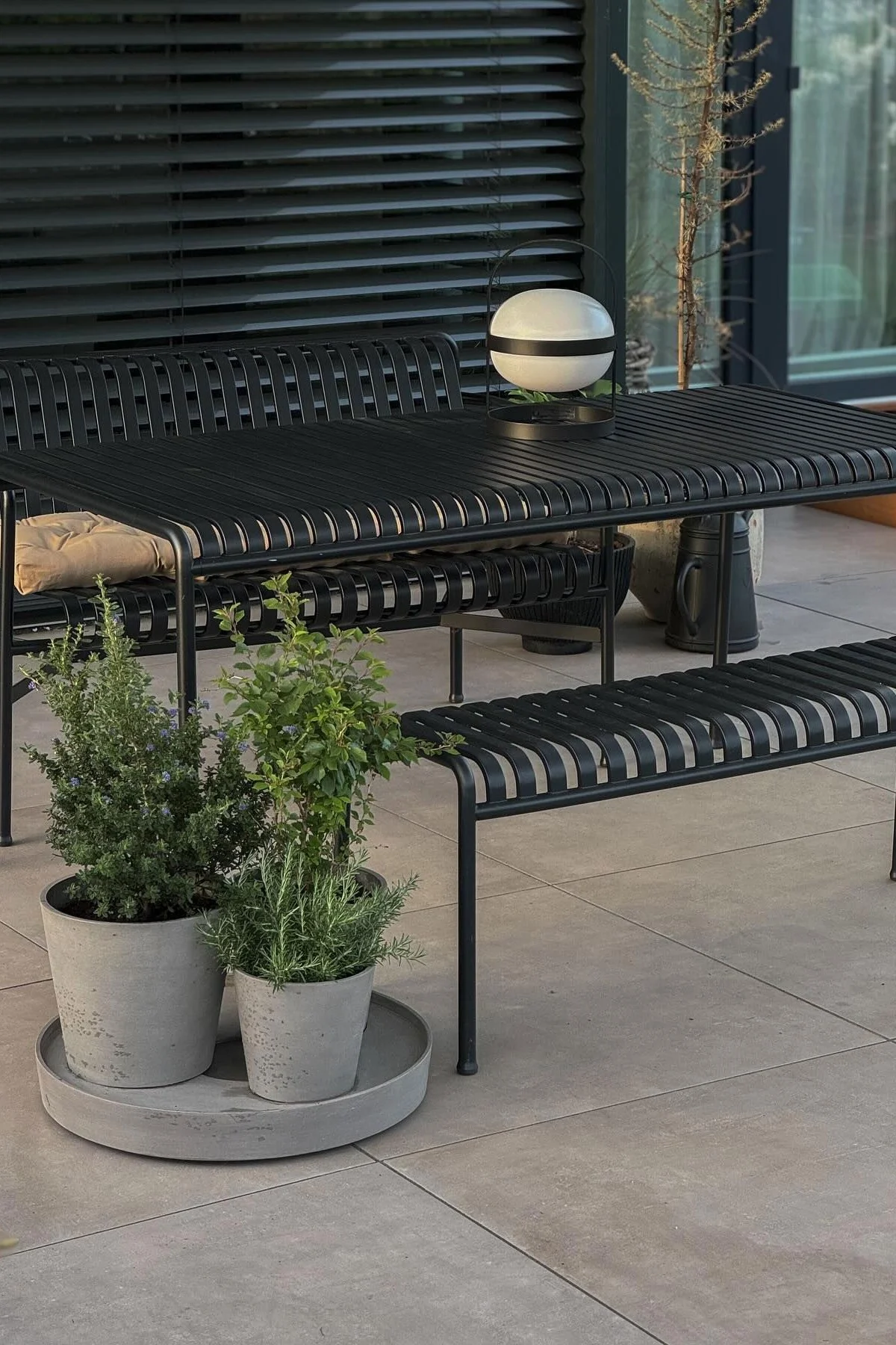 Svart utendørs spisestuesett i metall med lameller, sfærisk lykt og potteplanter på en uteplass.