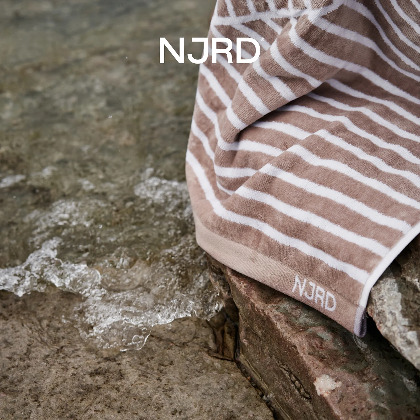 NJRD beige og hvitstripet håndkle på steiner ved krusende vann.