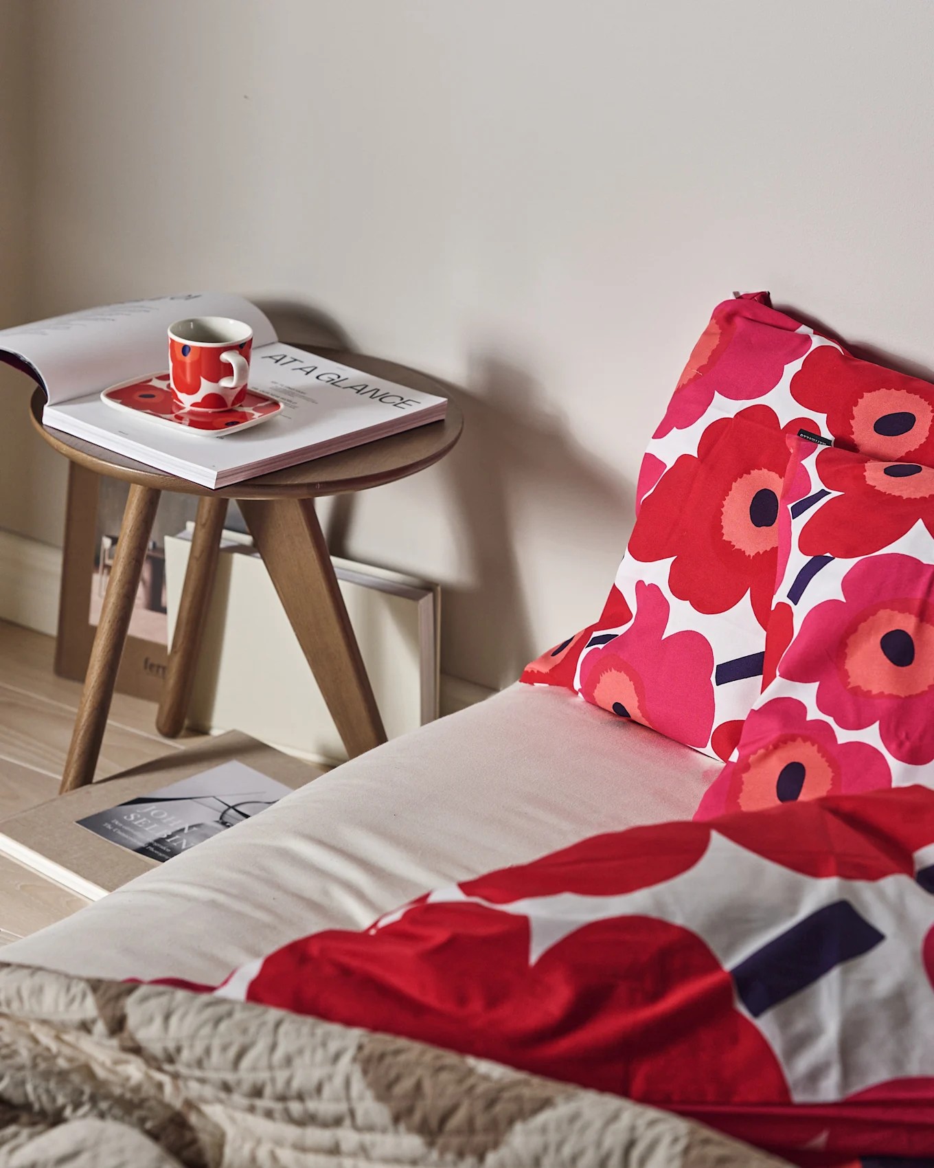 En seng med rødt og rosa blomstret sengetøy og et teksturert beige pledd. En rød kopp og åpen bok på et tresidebord.