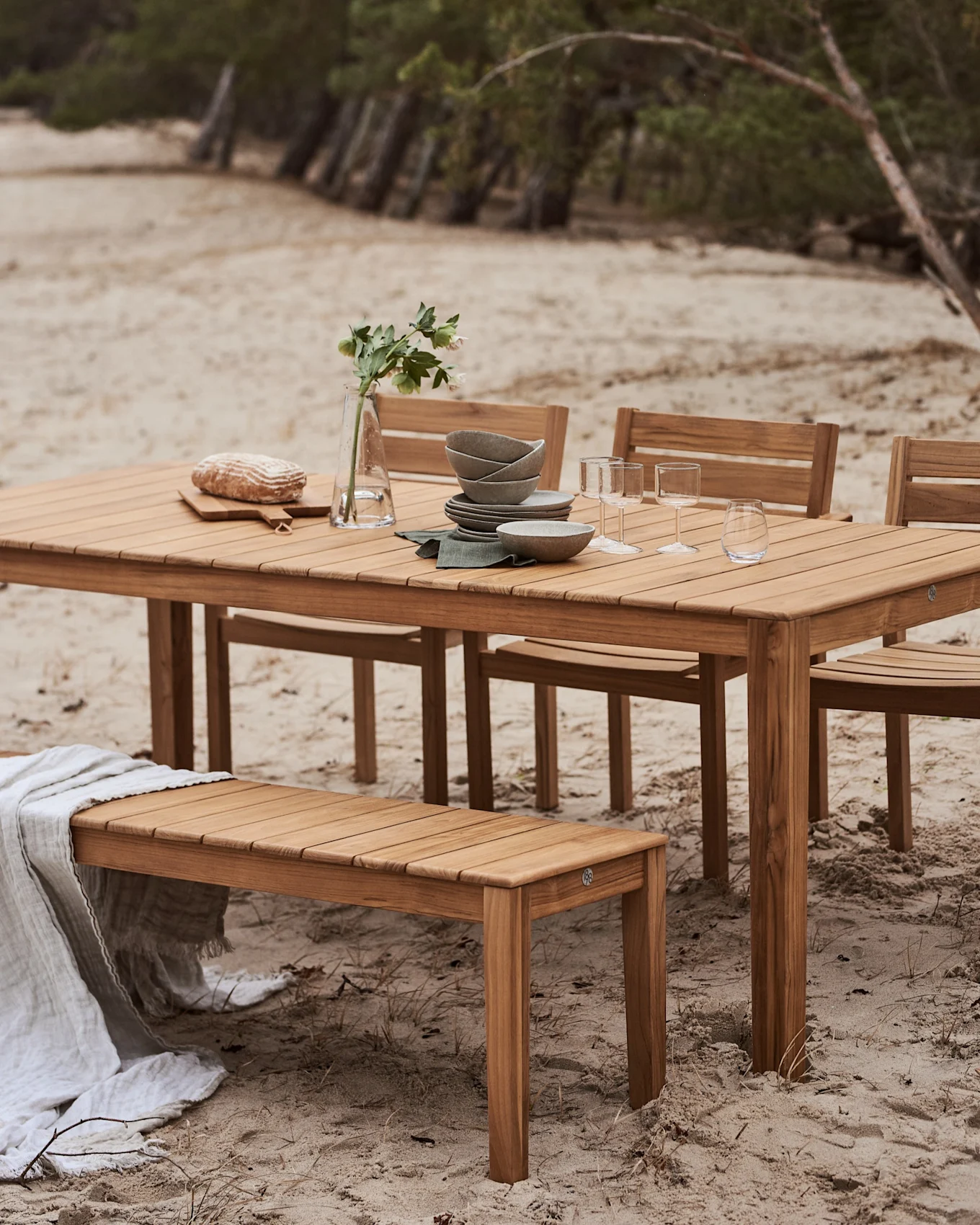 Utendørs spiseplass i tre med bord og benk på en sandstrand. Bordet er dekket med servise, glass, brød og en vase med blomster.