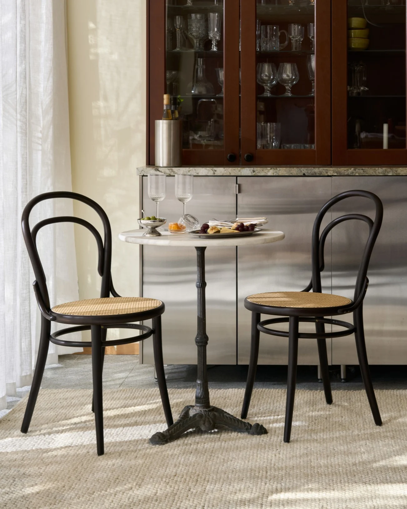 Et bistrobord med to mørke tre- og rottingstoler, snacks og vinglass, foran et skap med glassvarer.