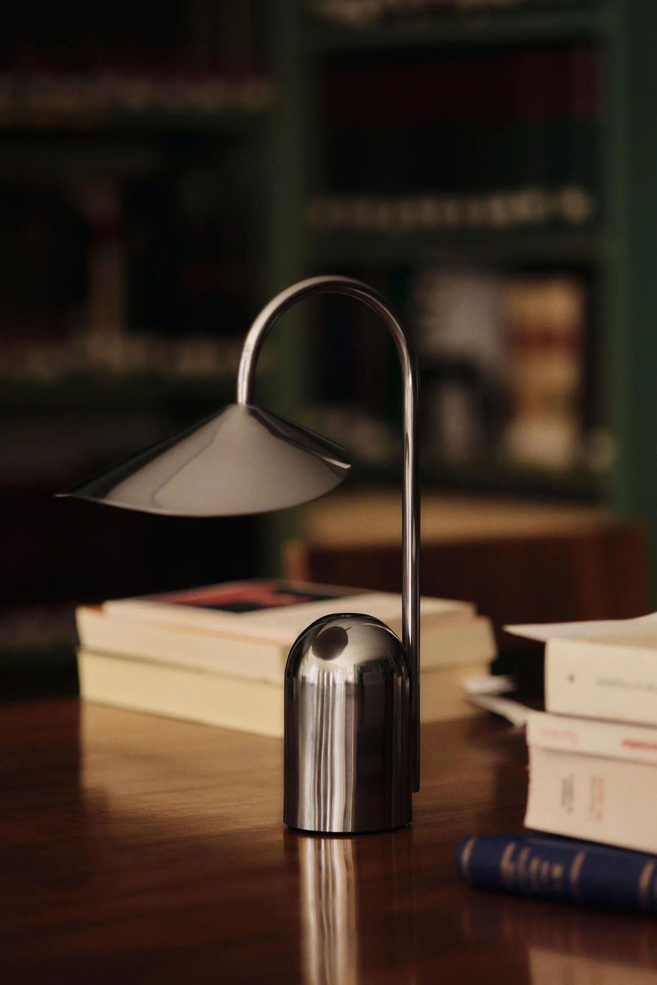En moderne sølv bordlampe med buet stamme og konisk skjerm står på et trebord med uskarpe bøker i bakgrunnen.