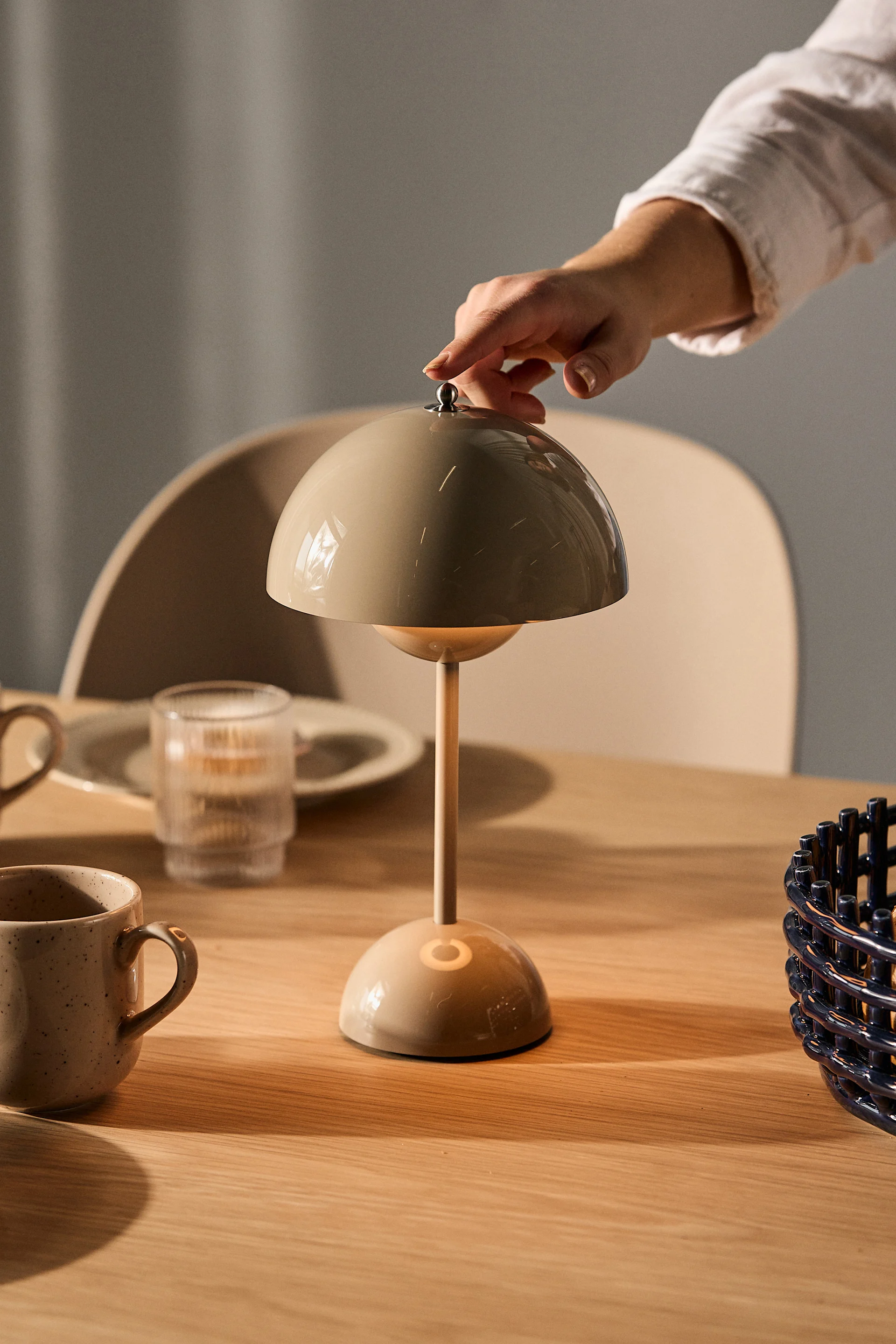 En hånd justerer en beige soppformet bordlampe på et trebord med krus og et glass.