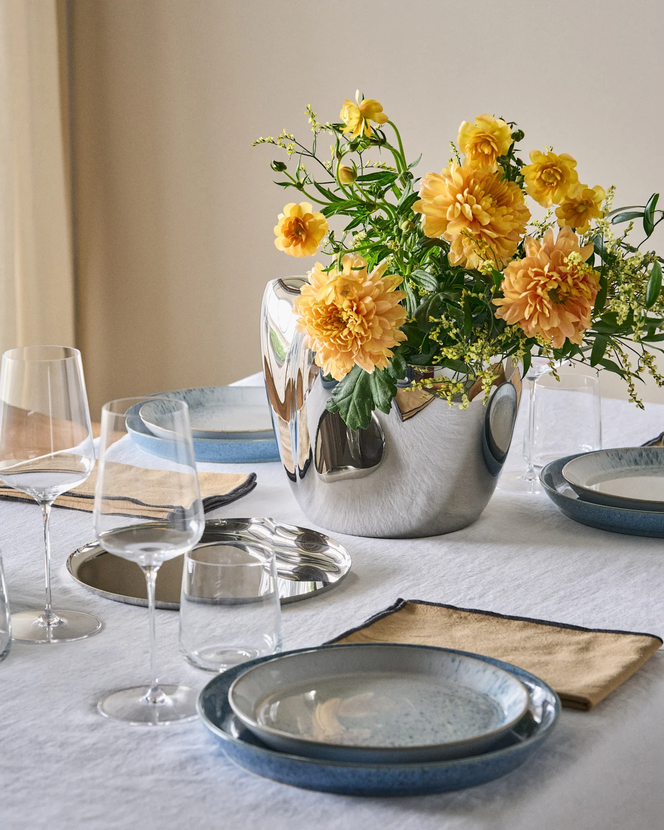 Et spisebord dekket med blå tallerkener, beige servietter, klare glass og en sølvvase med oransje-gule blomster på en hvit duk.