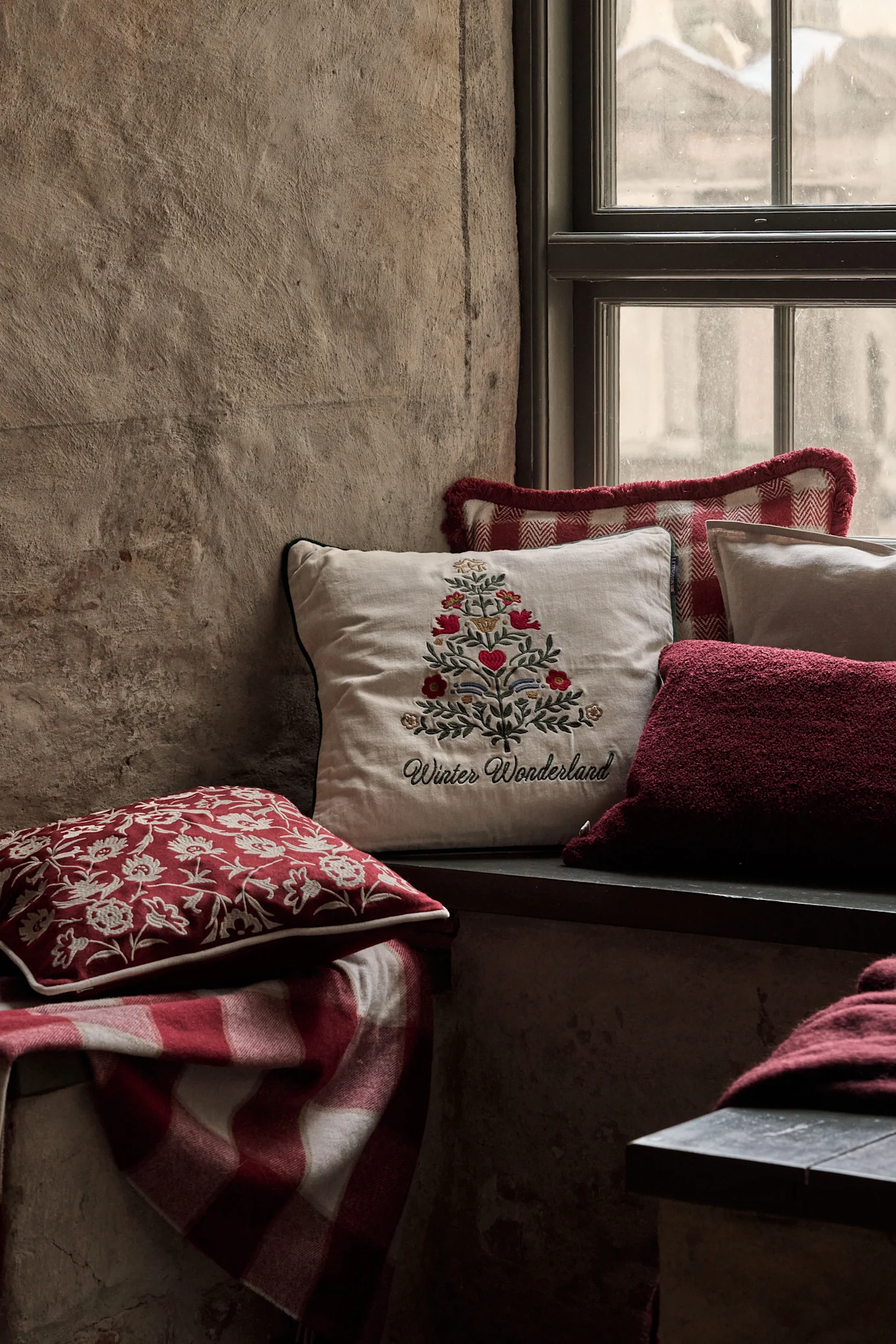Koselig vindusnisje med festlige puter, inkludert en med brodert juletre og 'Winter Wonderland', og et rødt rutete teppe.