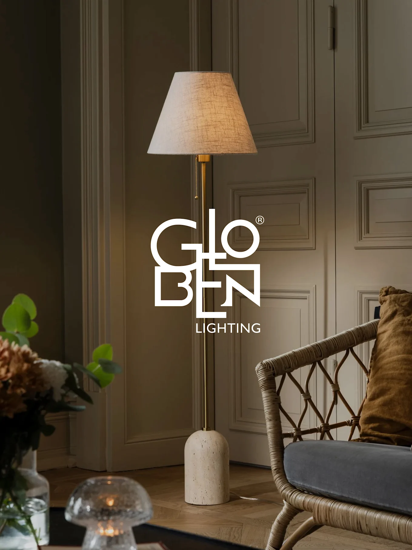 En gulvlampe med beige skjerm, messingstativ og avrundet steinbase står ved siden av en kurvstol.