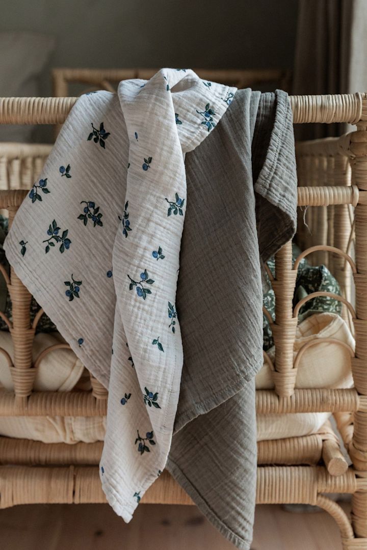 Blueberry Muslin-tepper fra Garbo&Friends, vist her hengende over en sengegavl, er allsidige tepper som kan brukes til mange ting, noe som gjør dem til perfekte babyshower-gaver.