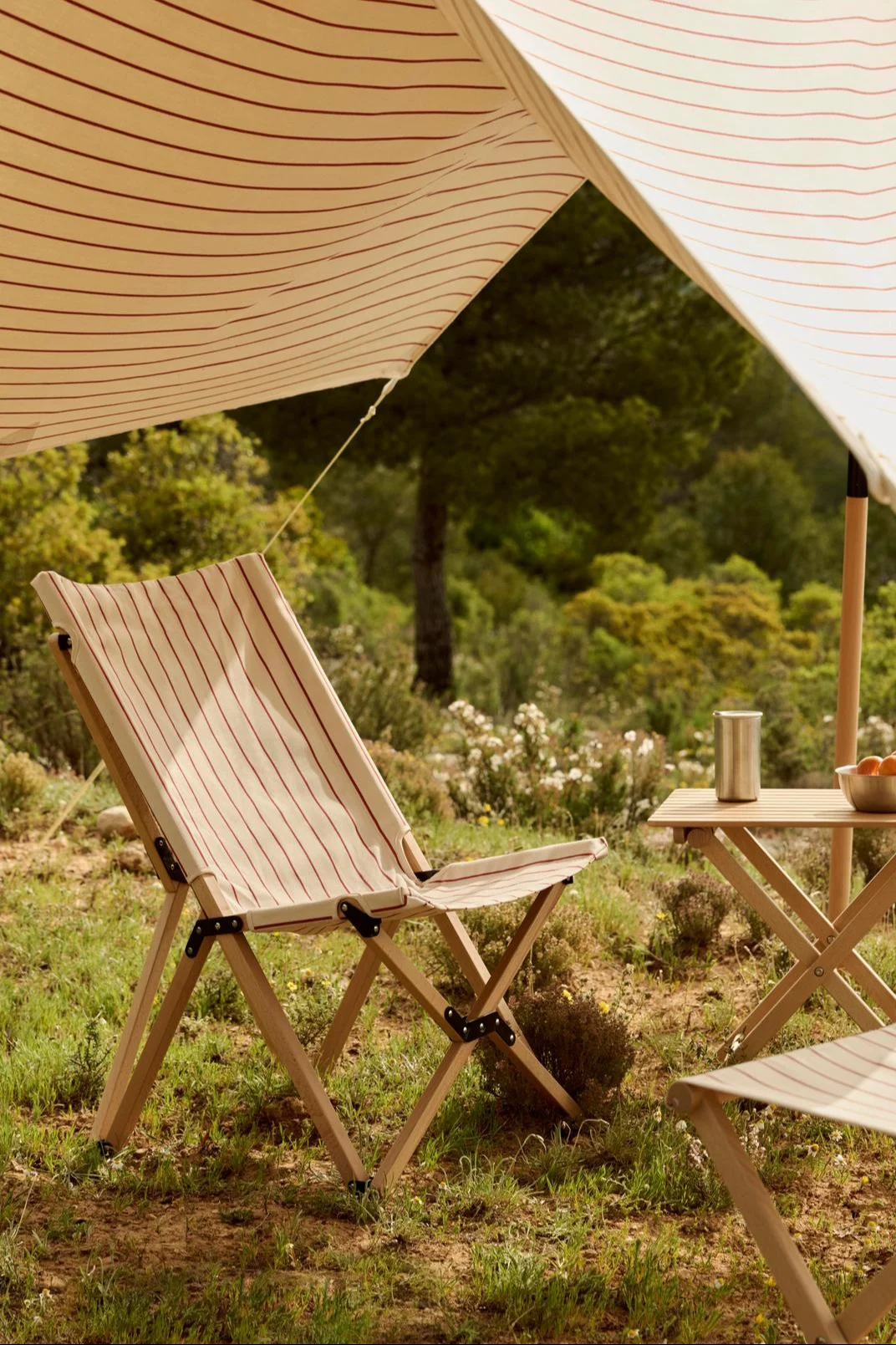 En stripete markise skygger en stripete sammenleggbar stol og et bord med termos og appelsiner i en naturlig utendørs setting.