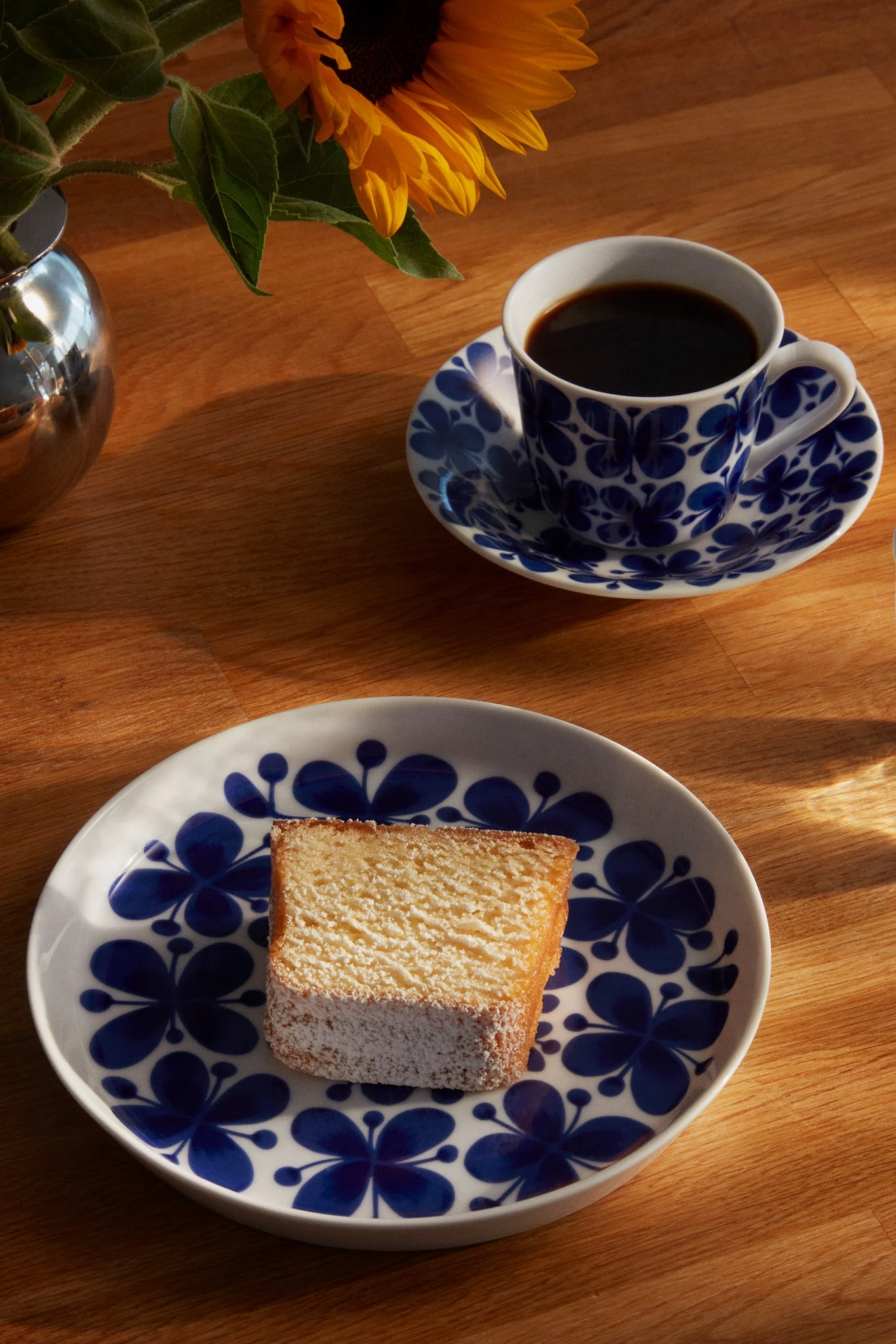Pulverkake på en blå blomstertallerken, svart kaffe i en matchende kopp og en solsikke i en sølvvase på tre.