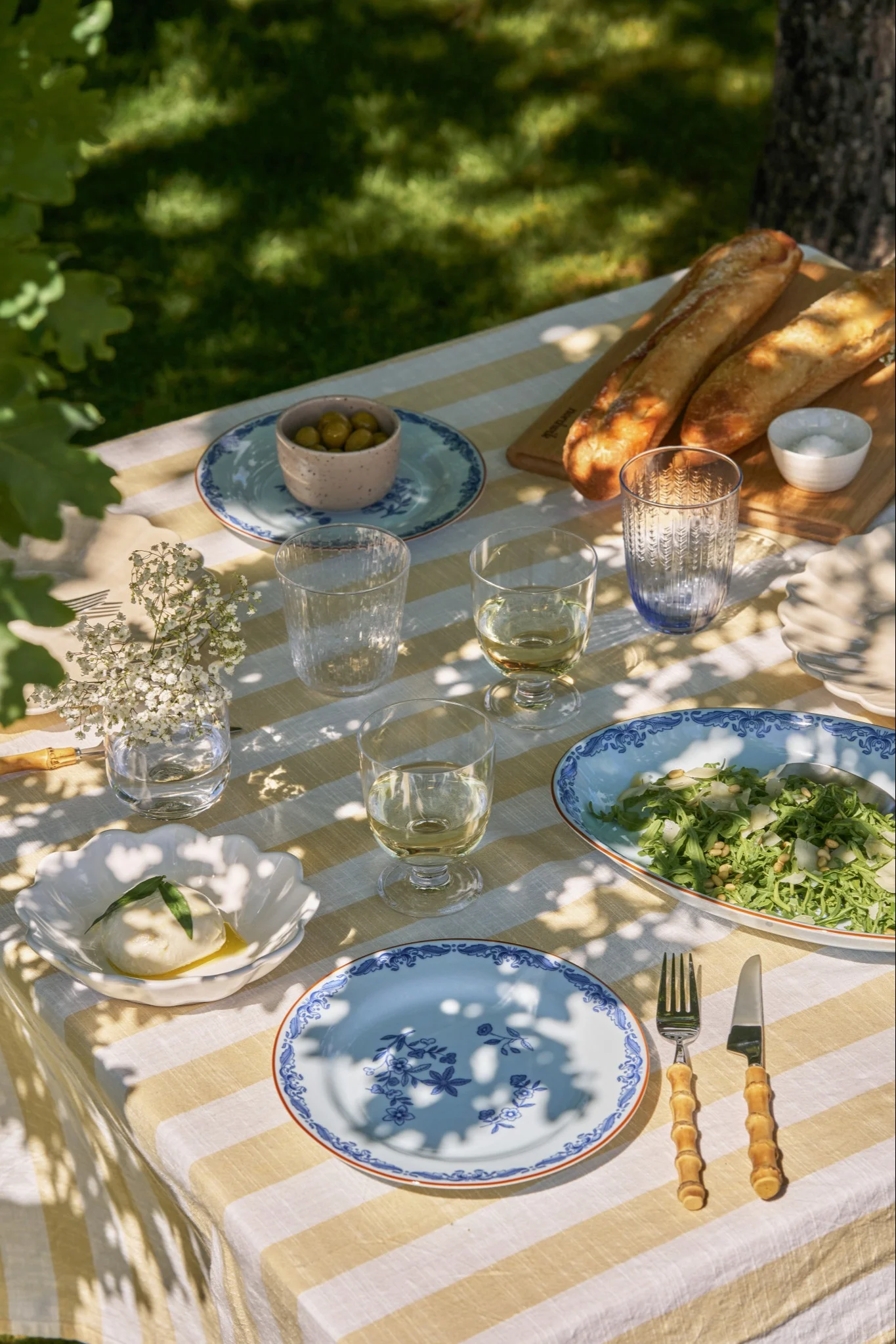 Et utendørs piknikbord dekket med en gul og hvit stripet duk, burrata, salat, baguetter, oliven og blåmønstrede tallerkener.
