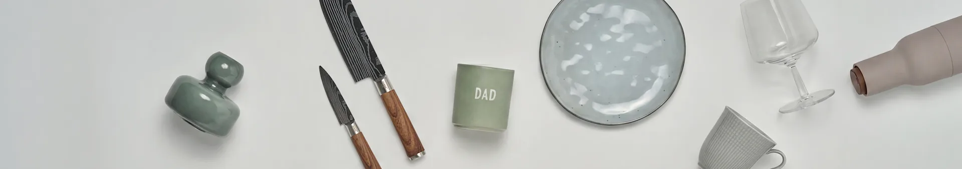 Oversiktsbilde av diverse husholdningsartikler: en grønn abstrakt vase, to kniver, et 'DAD'-lys, en flekkete tallerken, et vinglass, en grå kopp og en beige flaske på hvitt.