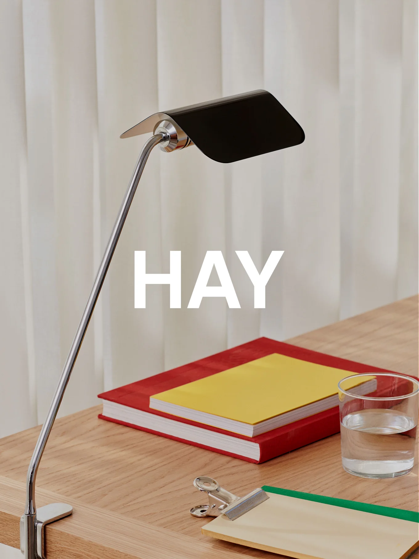 En krom bordlampe med svart skjerm belyser et treskrivebord med to bøker, et glass vann og et skrivebrett.