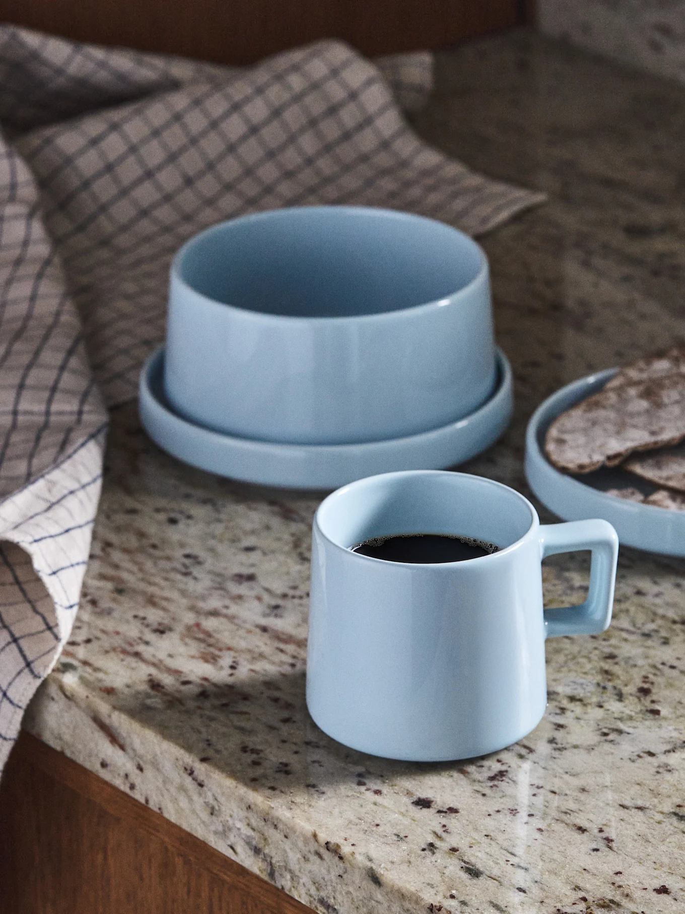 Lys blå keramisk kaffekopp, bolle og tallerken med flatbrød på en granittbenk.