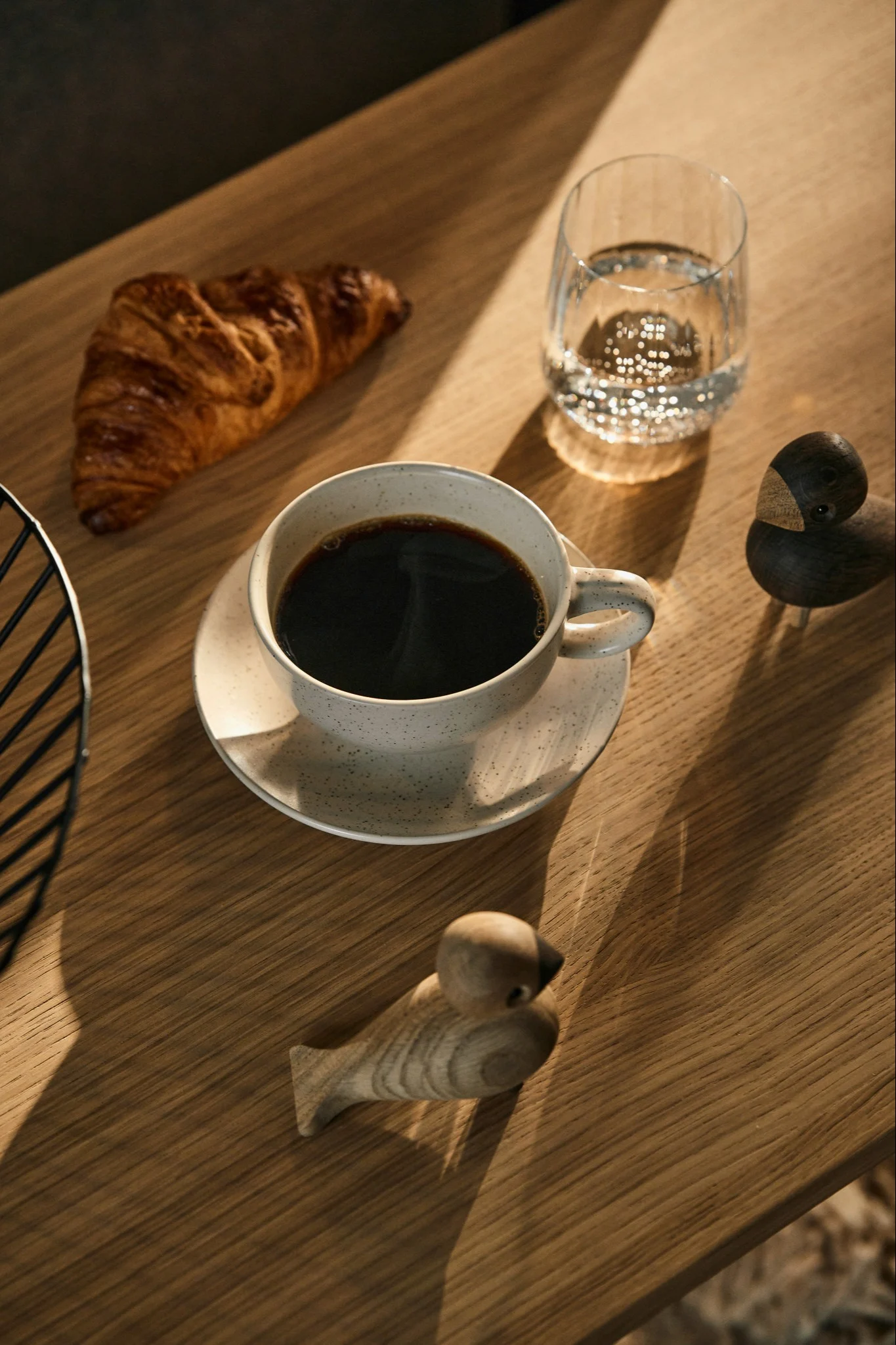 Et frokostbord med kaffe, en croissant, vann og to trefuglfigurer på et solrikt trebord.
