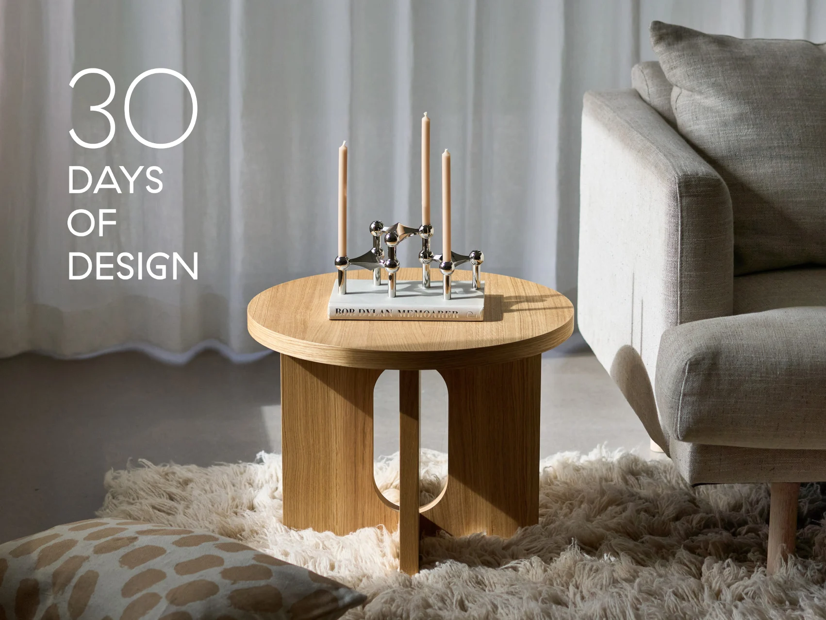 Moderne stue med rundt sidebord i tre, sølv lysestake med tre lys, en bok og en beige sofa. Tekst: '30 DAGER MED DESIGN'.