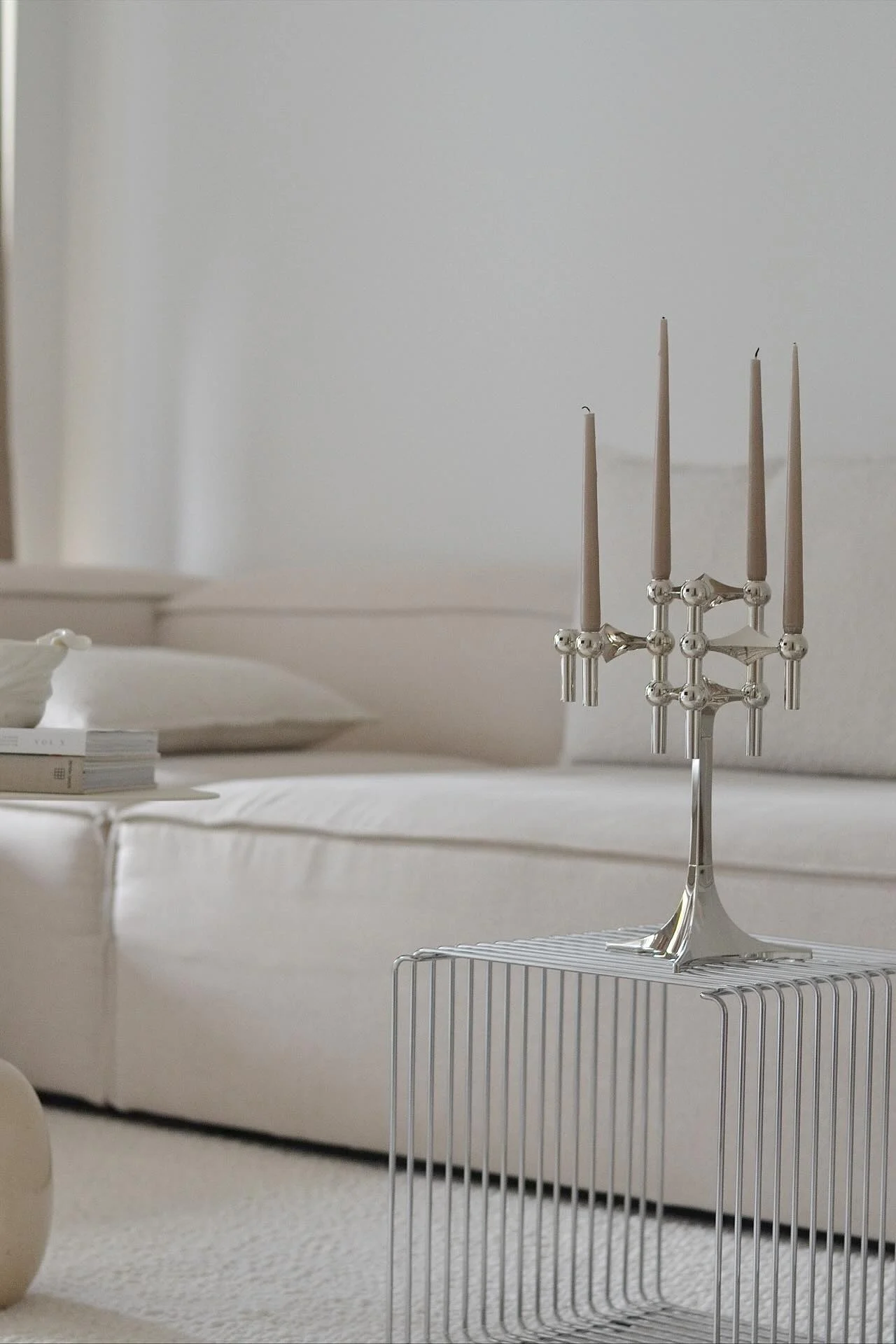 En sølv kandelaber med fem lysebrune stearinlys står på et minimalistisk sidebord i metall. En lys sofa er i bakgrunnen.