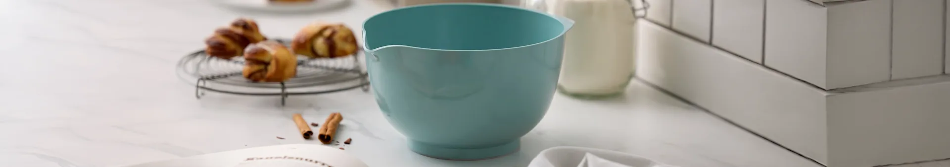 Lyseblå miksebolle, bakverk på rist, melkeflaske og kokebok på marmorbenkeplate.