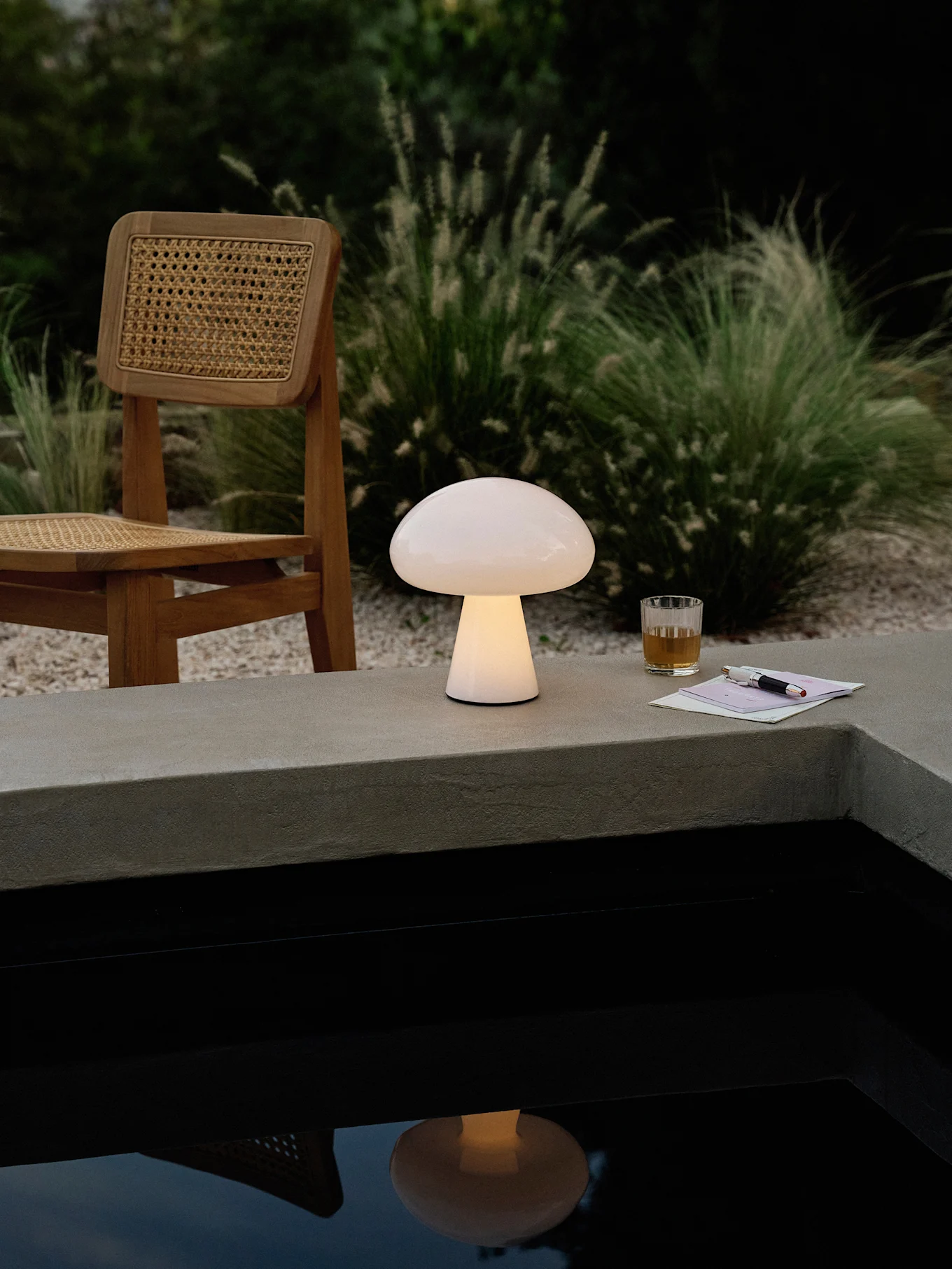 En glødende soppformet lampe på en betongkant ved siden av et basseng, med en trestol, drikke og penn.