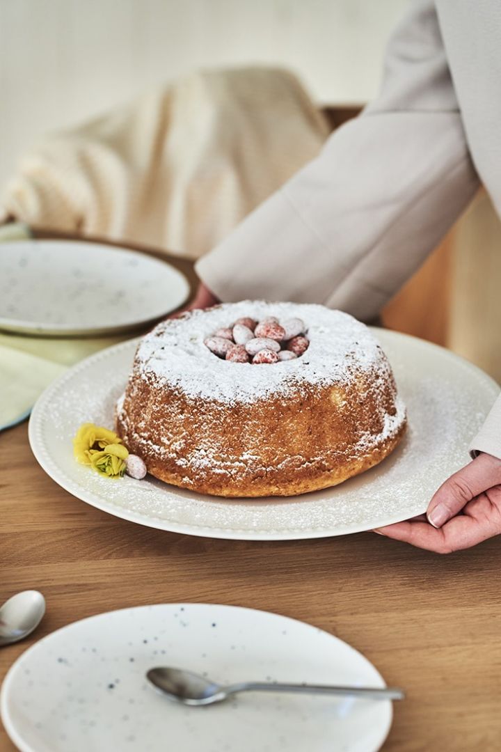 Årets påskedessert, et sukkerbrød, serveres på et hvitt serveringsfat fra Scandi Living med et påskeegg på og en gul blomst som pynt.