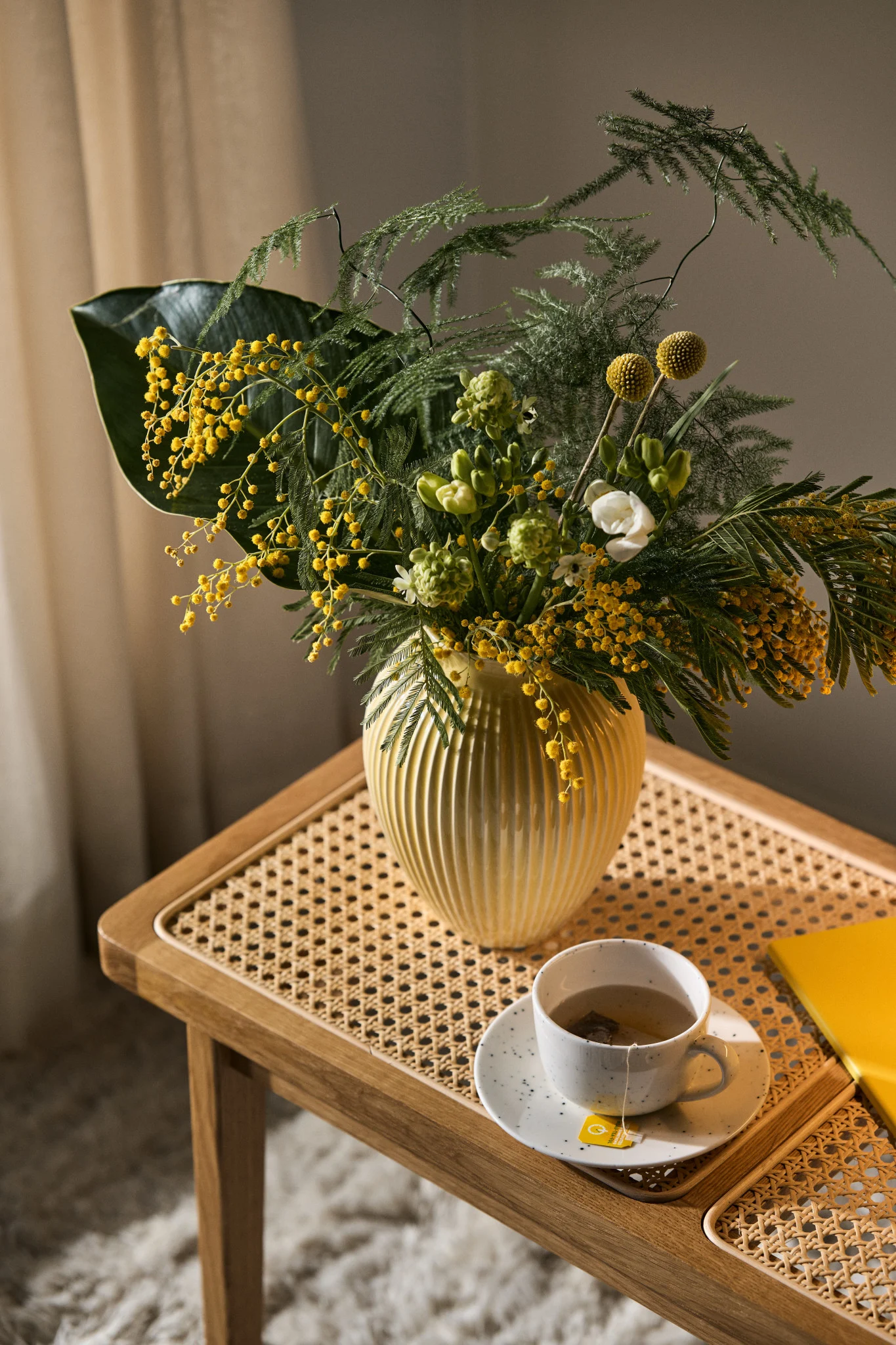 På en trebenk står en kopp kaffe og en vakkert arrangert blomsterbukett med snittblomster i en riflet, gul blomstervase fra Knabstrup.