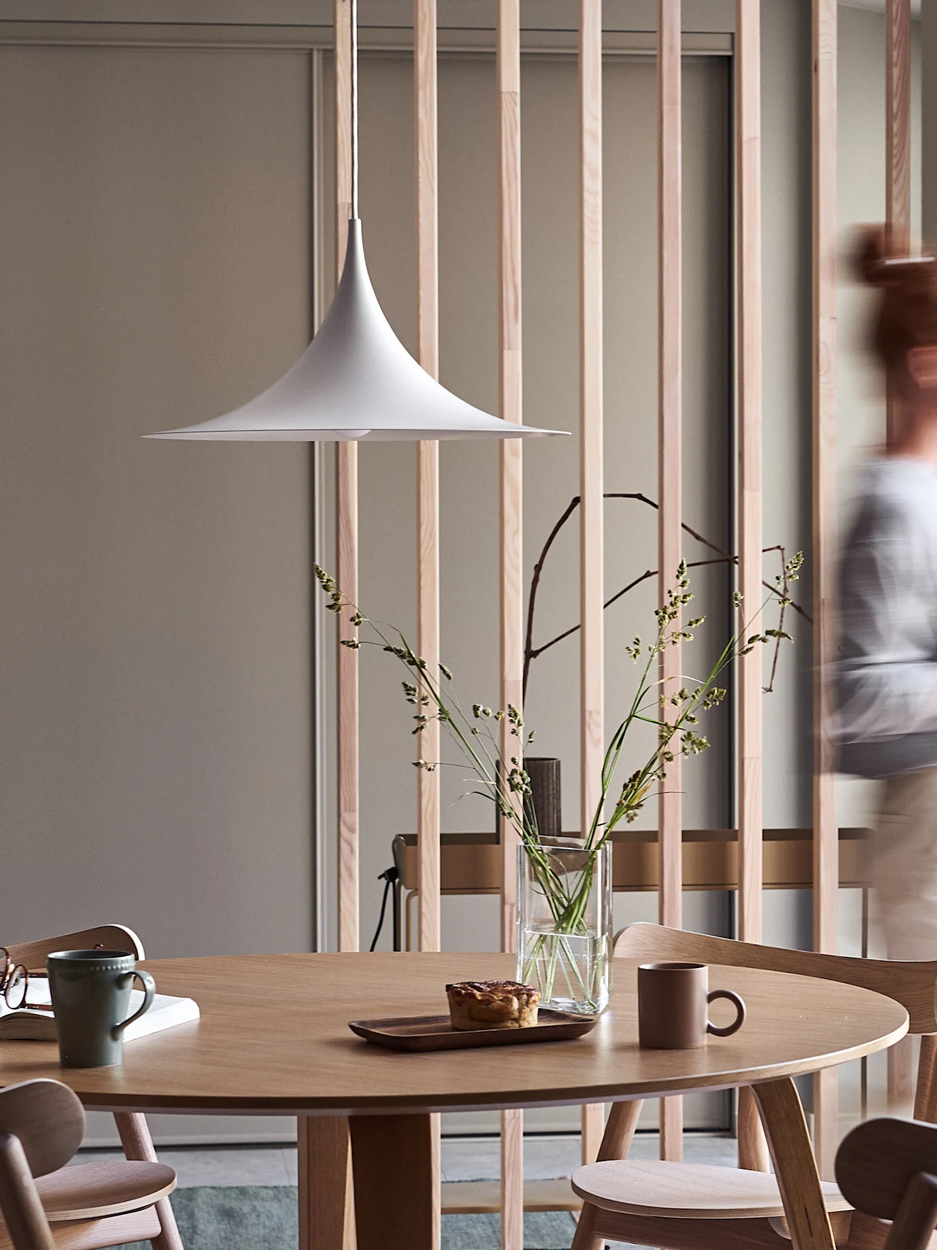 Moderne spiseområde med rundt trebord, hvit pendellampe, vase med gress, bakverk og krus, med en uskarp person som går forbi.