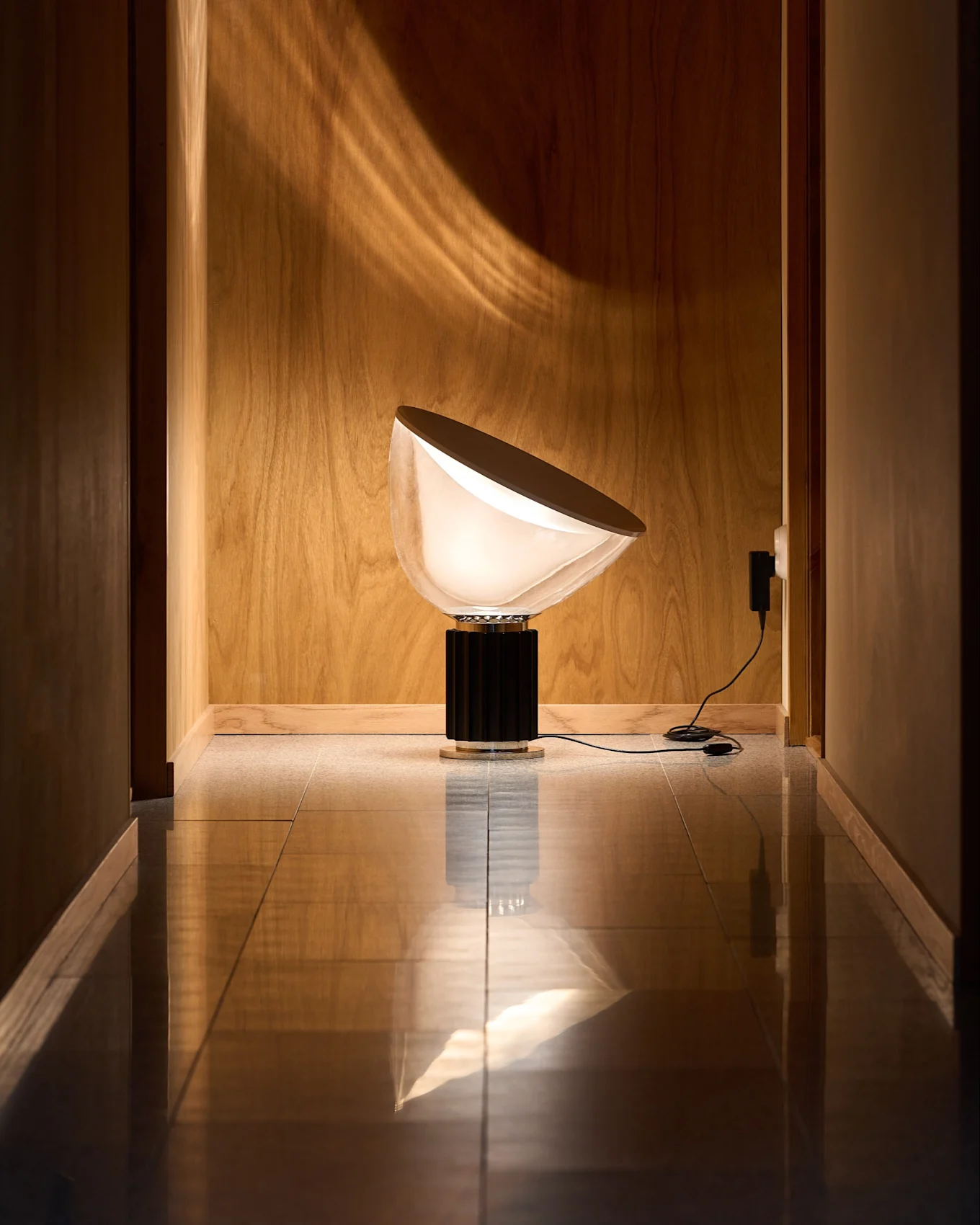 Moderne lampe med halvrund, hvit skjerm og svart riflet base i trepanelert gang med reflekterende gulv. Lys danner buet mønster på veggen.