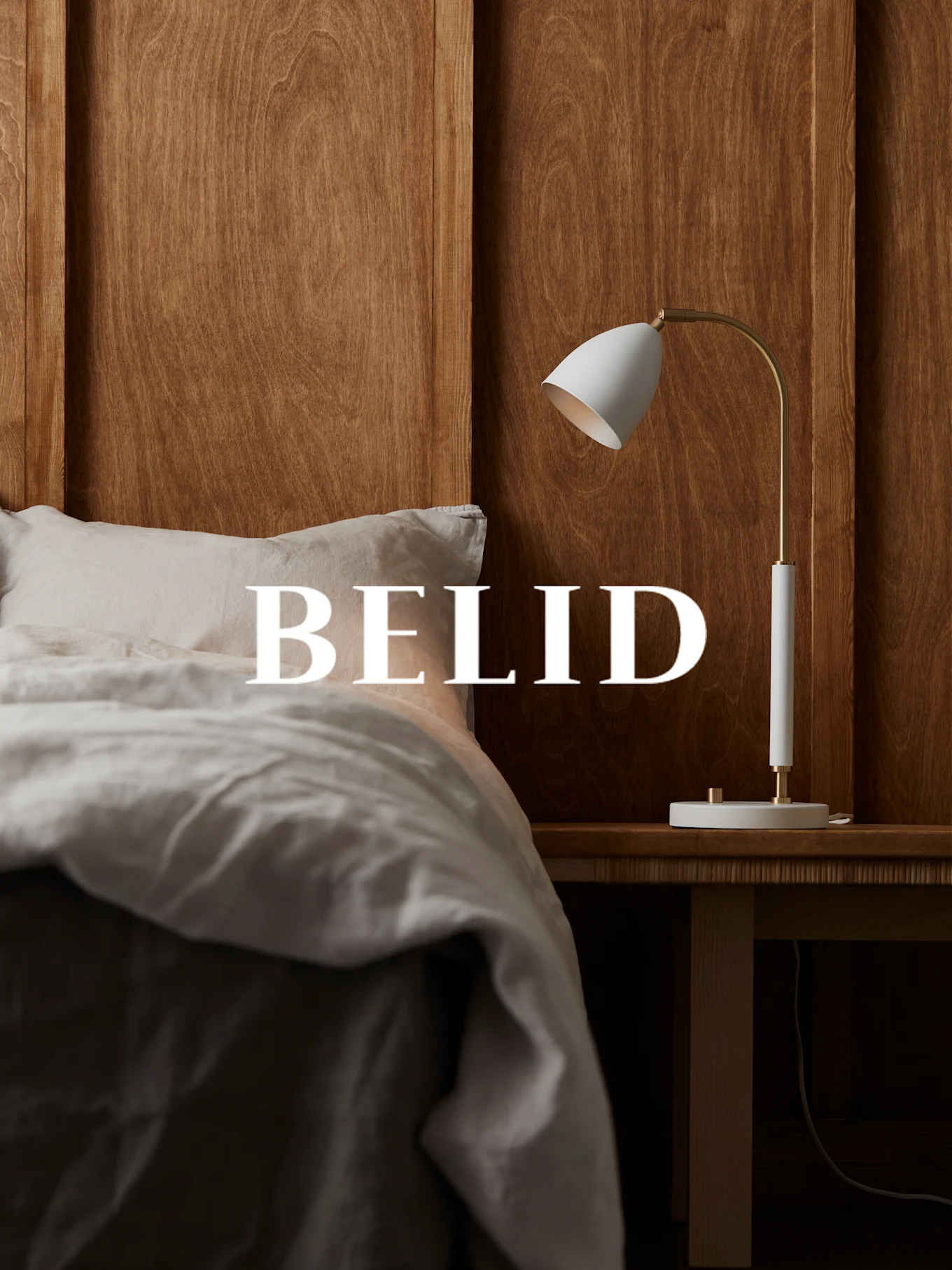 Hvit og messinglampe på et nattbord i tre, med 'BELID'-tekst over en seng med linsengetøy.