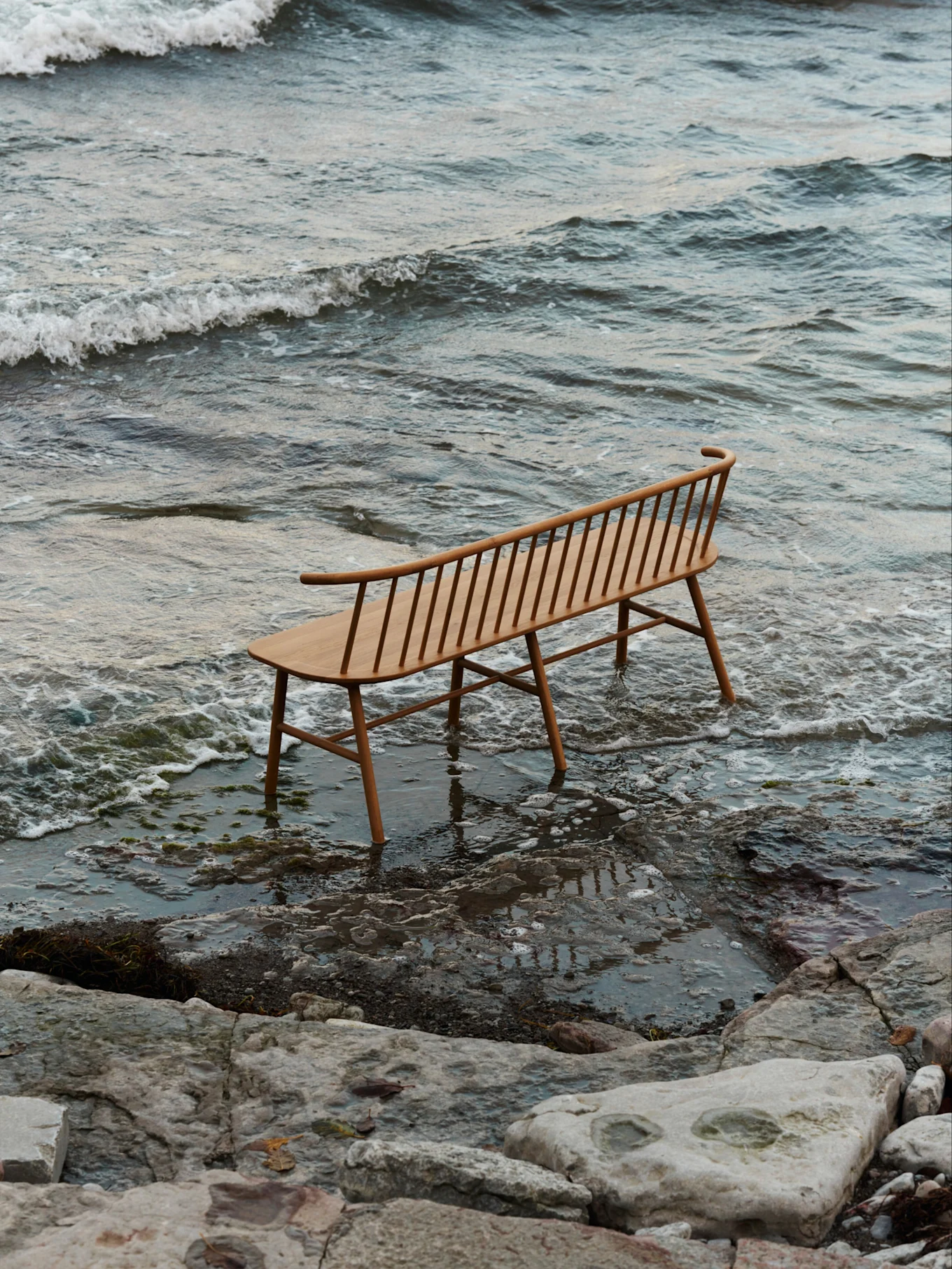 En lys trebenk med spiler og buet ryggstøtte står i grunt vann på en steinstrand med milde bølger.