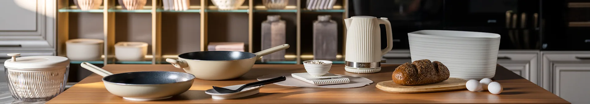 Kjøkkenbenk med beige stekepanner, en hvit riflet vannkoker, et brød med frø på et brett og egg. Moderne kjøkkenutstyr.