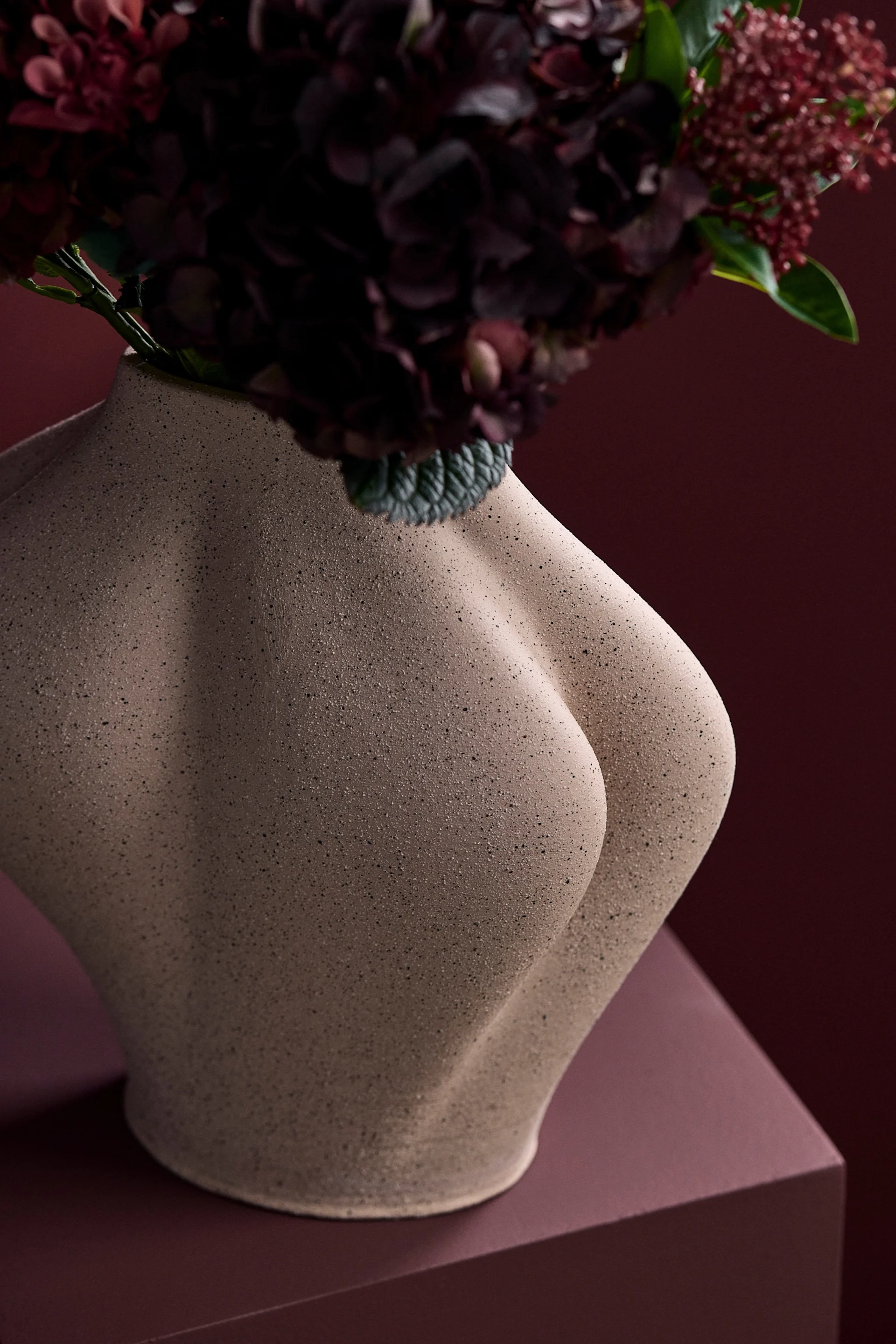 Skulpturell beige vase formet som en menneskelig torso, med mørkerøde blomster mot en matchende rød bakgrunn.