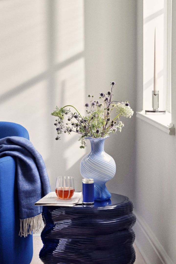 Mella vase fra Broste Copenhagen i blå farge med stripet mønster står på et sidebord ved siden av en blå sofa, og inneholder en bukett blomster.