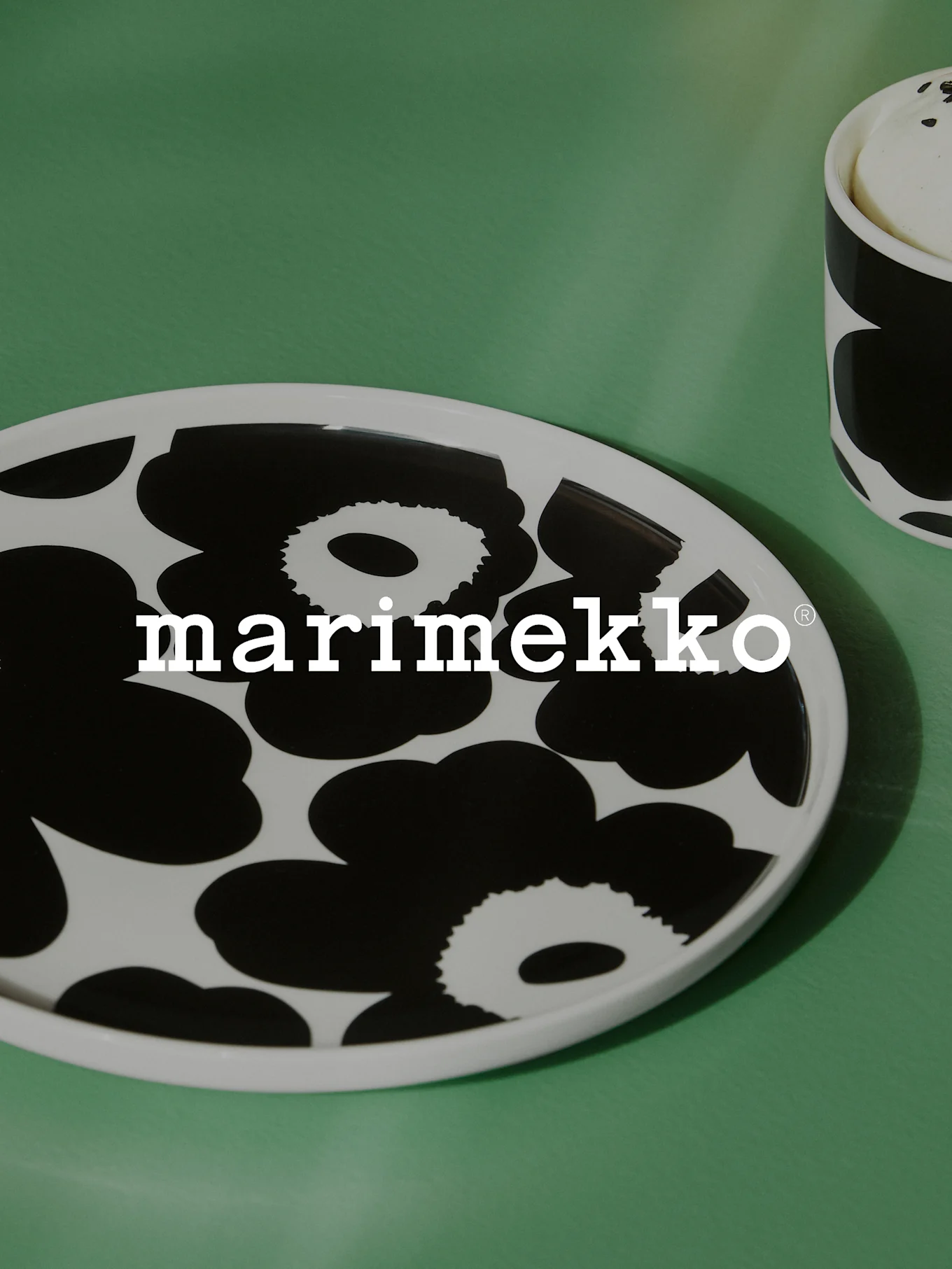Hvit oval tallerken med svart blomstermønster og Marimekko-logo på en grønn overflate, med en delvis matchende kopp.