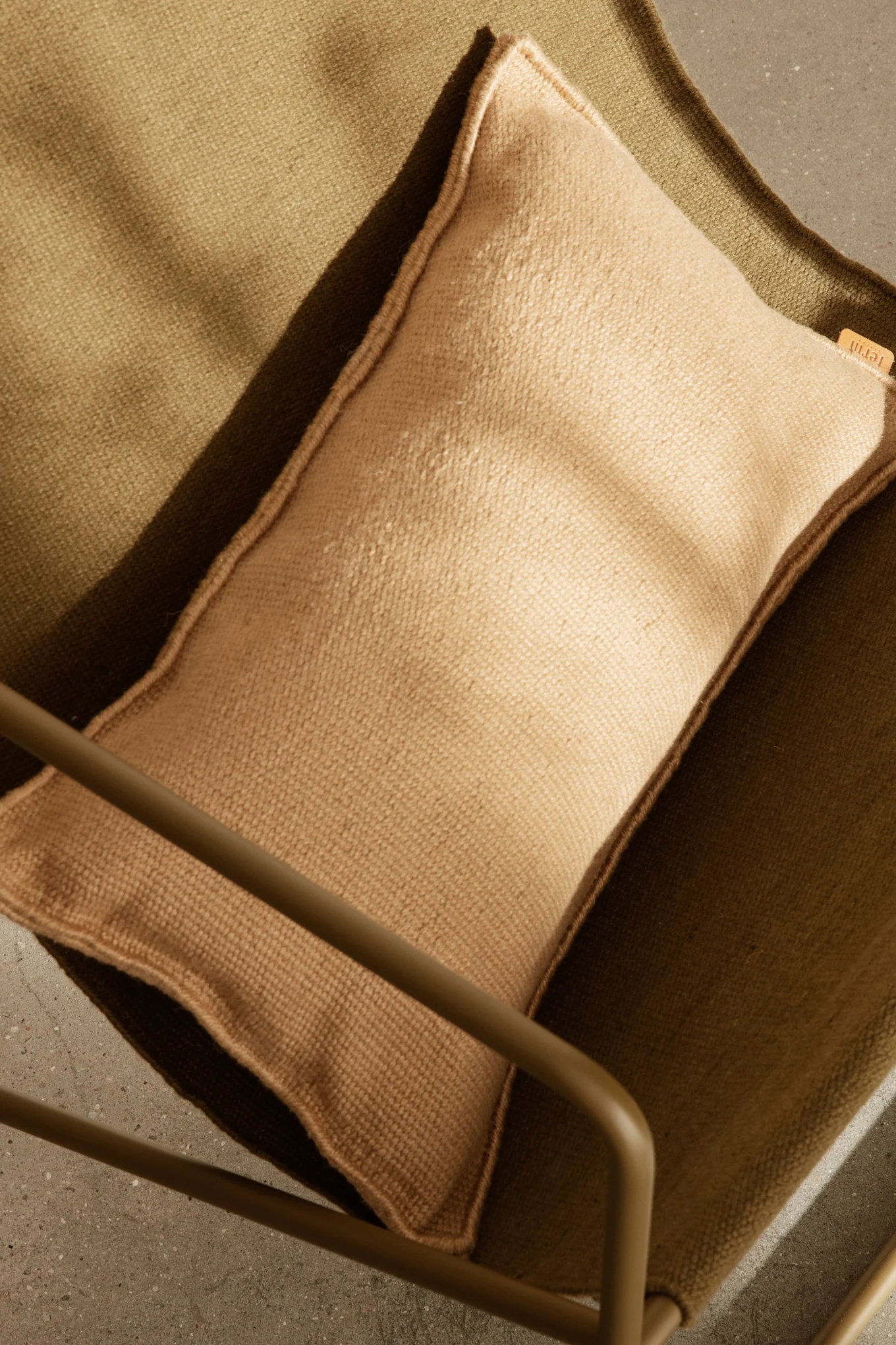 Ovenfra-bilde av en lysebrun pute på en mørk olivengrønn teksturert stoffstol, med del av en metallramme og betonggulv.