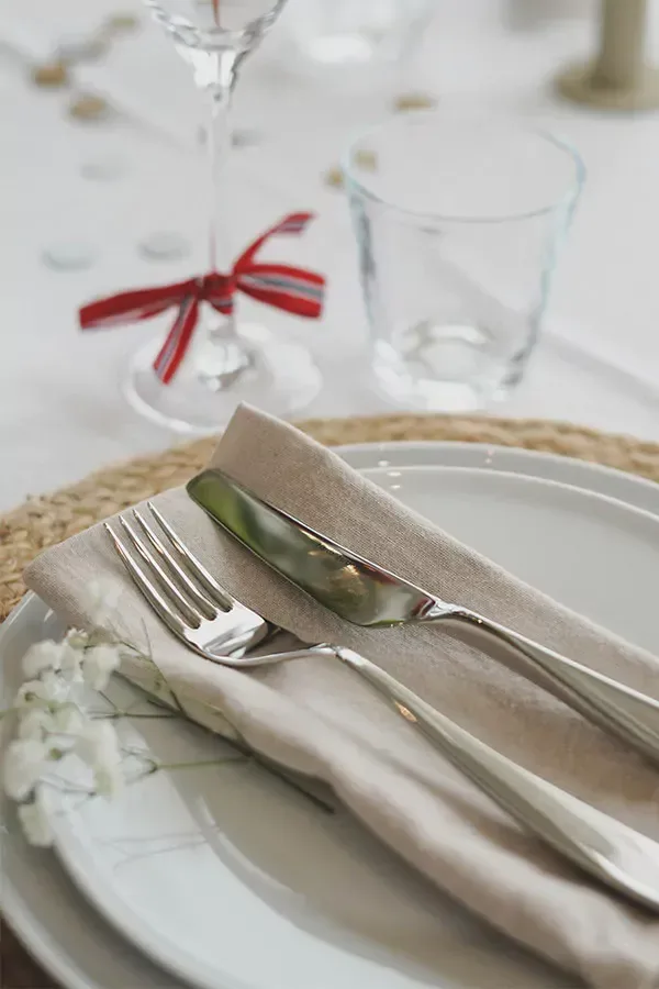 Elegant kuvert med hvite tallerkener, en beige serviett, sølvbestikk og et vinglass med rødt bånd.