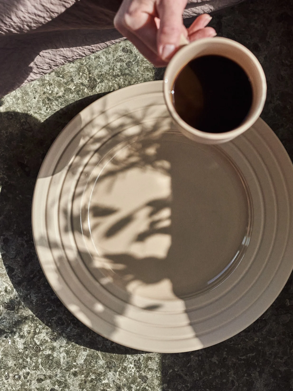 Hånd holder kaffekopp over beige tallerken med bladskygger på et flekkete bord.