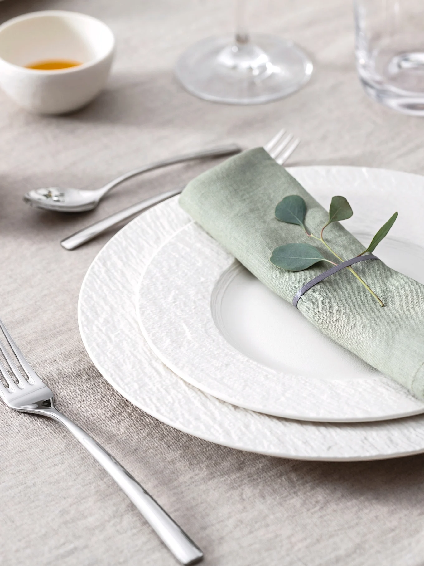 Elegant borddekking med lysegrønn linserviett, eukalyptusblader, hvite teksturerte tallerkener og bestikk på en beige duk.