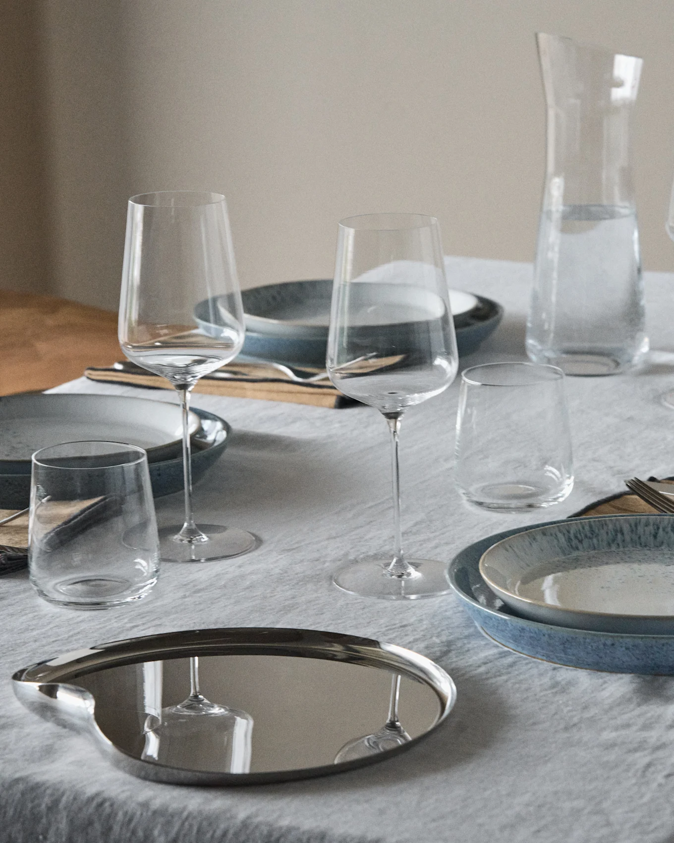 Et dekket bord med blå og flekkete tallerkener, vinglass, vannglass, en vannkaraffel og et reflekterende ovalt brett.